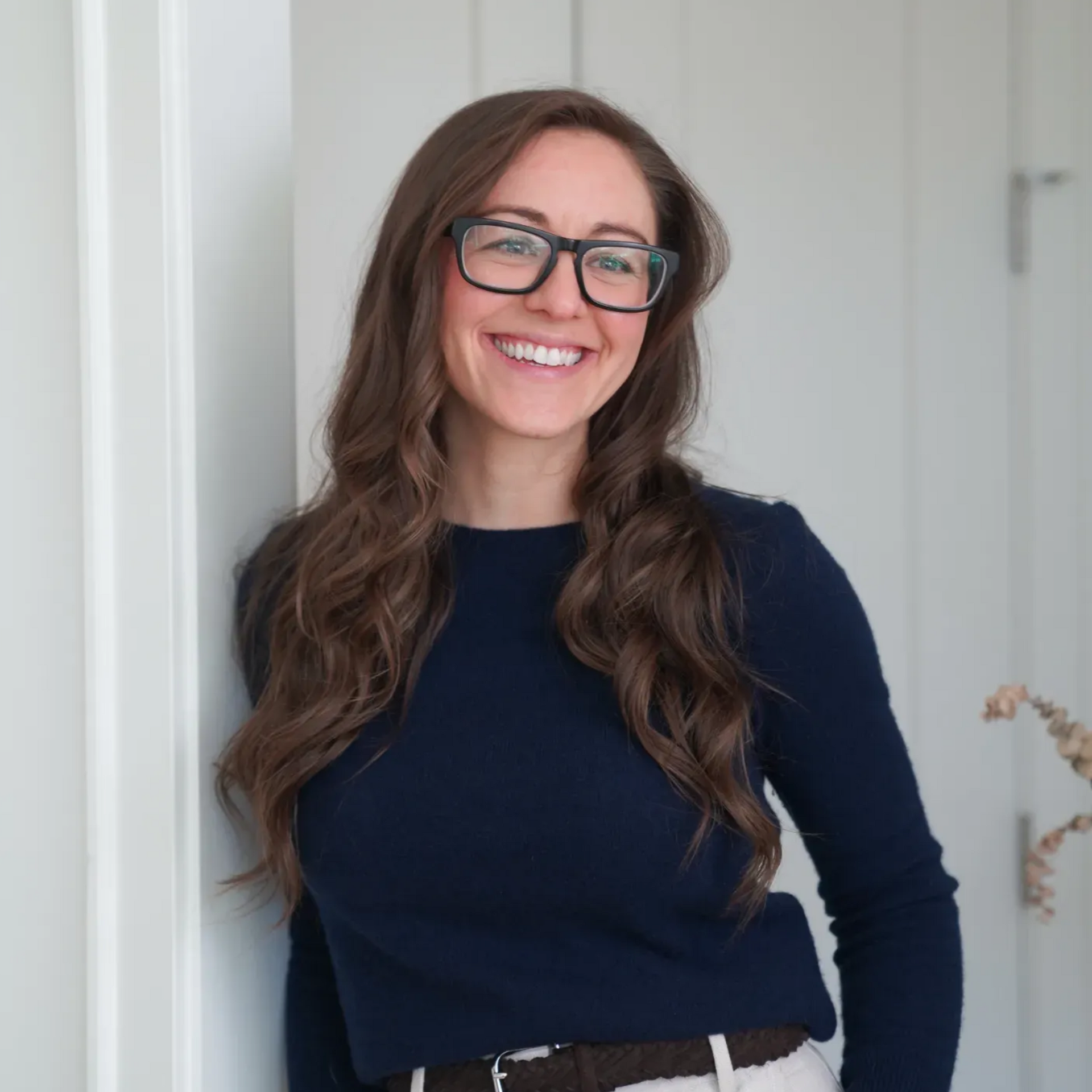 Smiling woman with glasses in a navy top, standing indoors by a light-colored wall