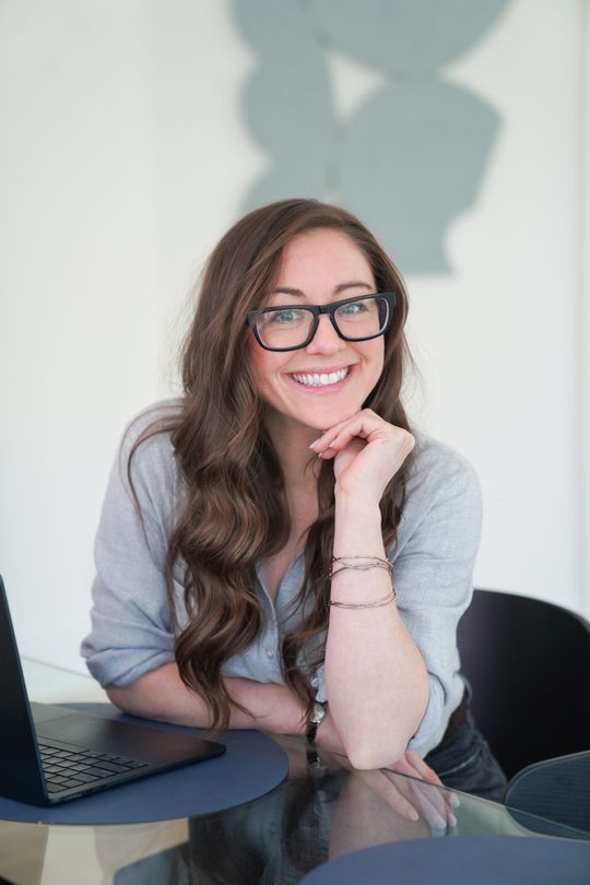 Smiling woman with glasses leaning on a table beside a laptop in a bright office
