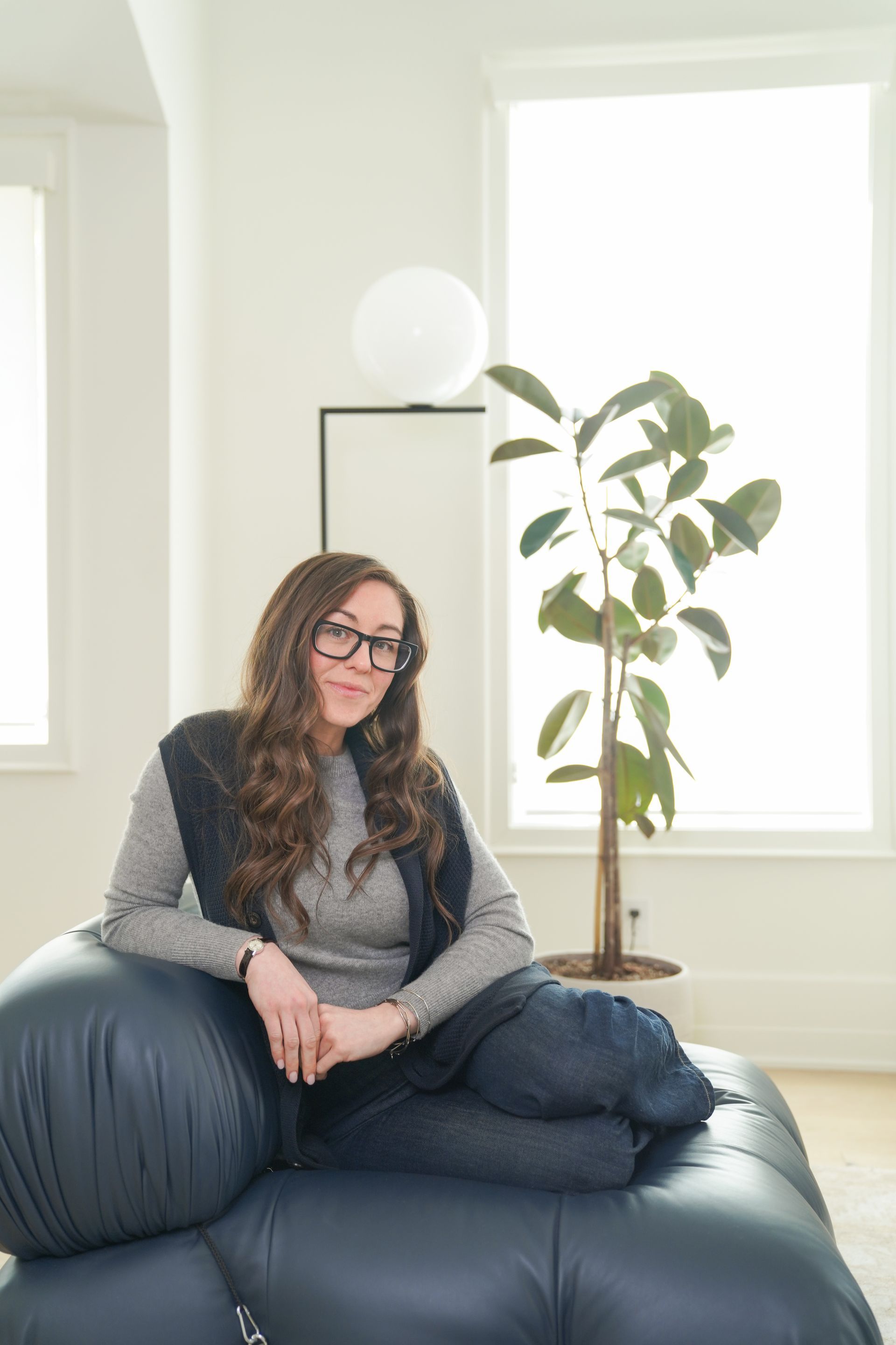 Woman seated on a dark blue sofa in a bright room with a potted plant and lamp nearby