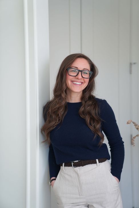 Smiling person in glasses and navy sweater, leaning against a doorway in light pants.