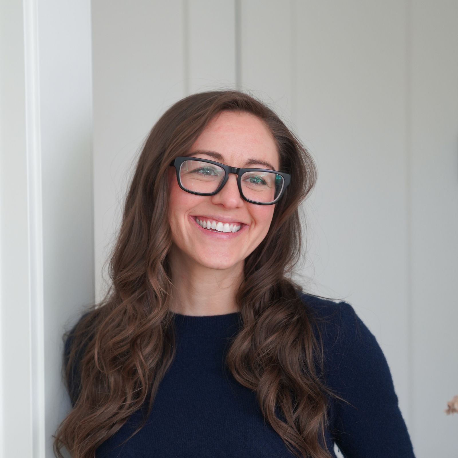 Smiling woman with glasses and long wavy brown hair against a light gray wall