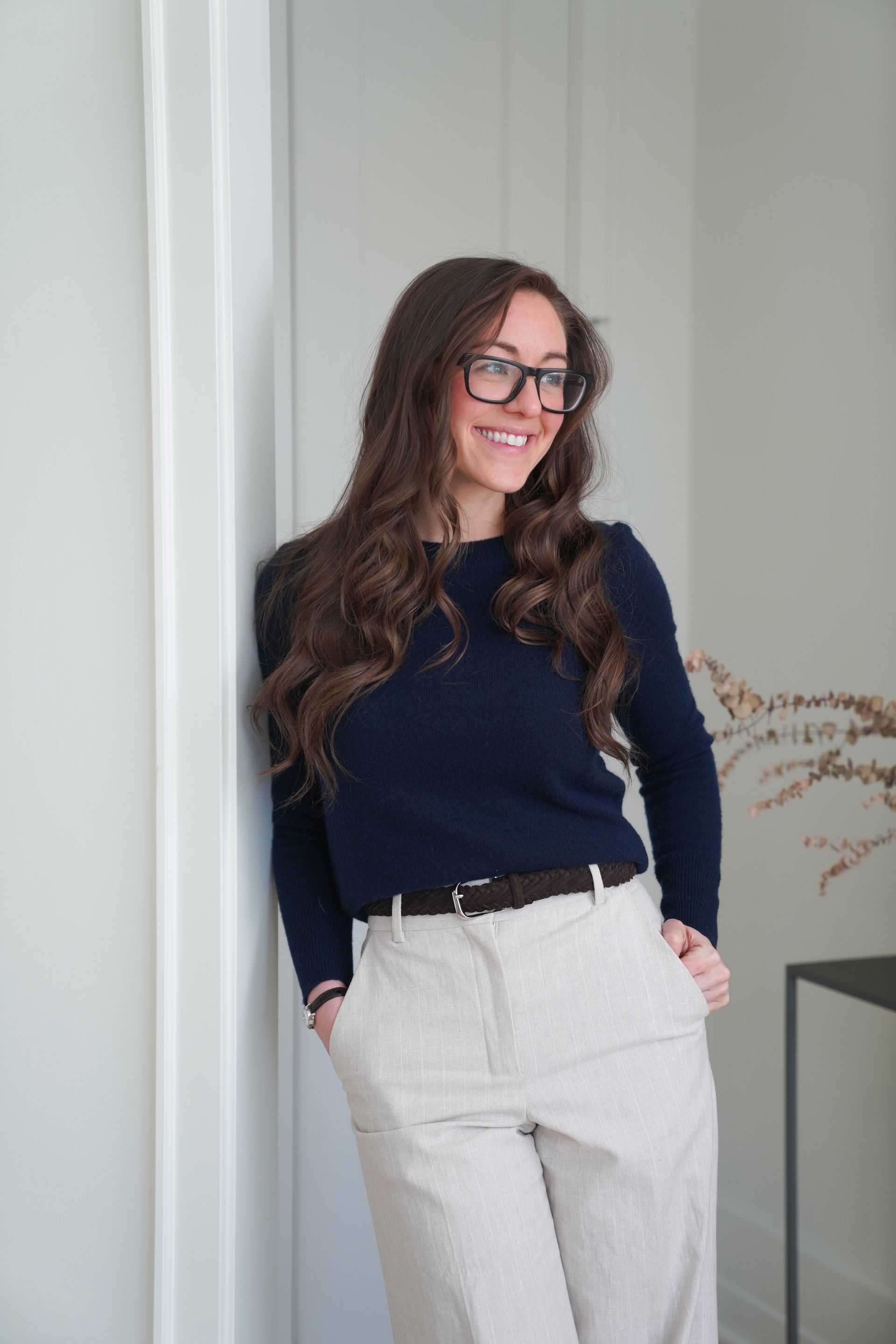 Person standing by a wall, smiling, wearing a navy top and light pants, with hands in pockets.