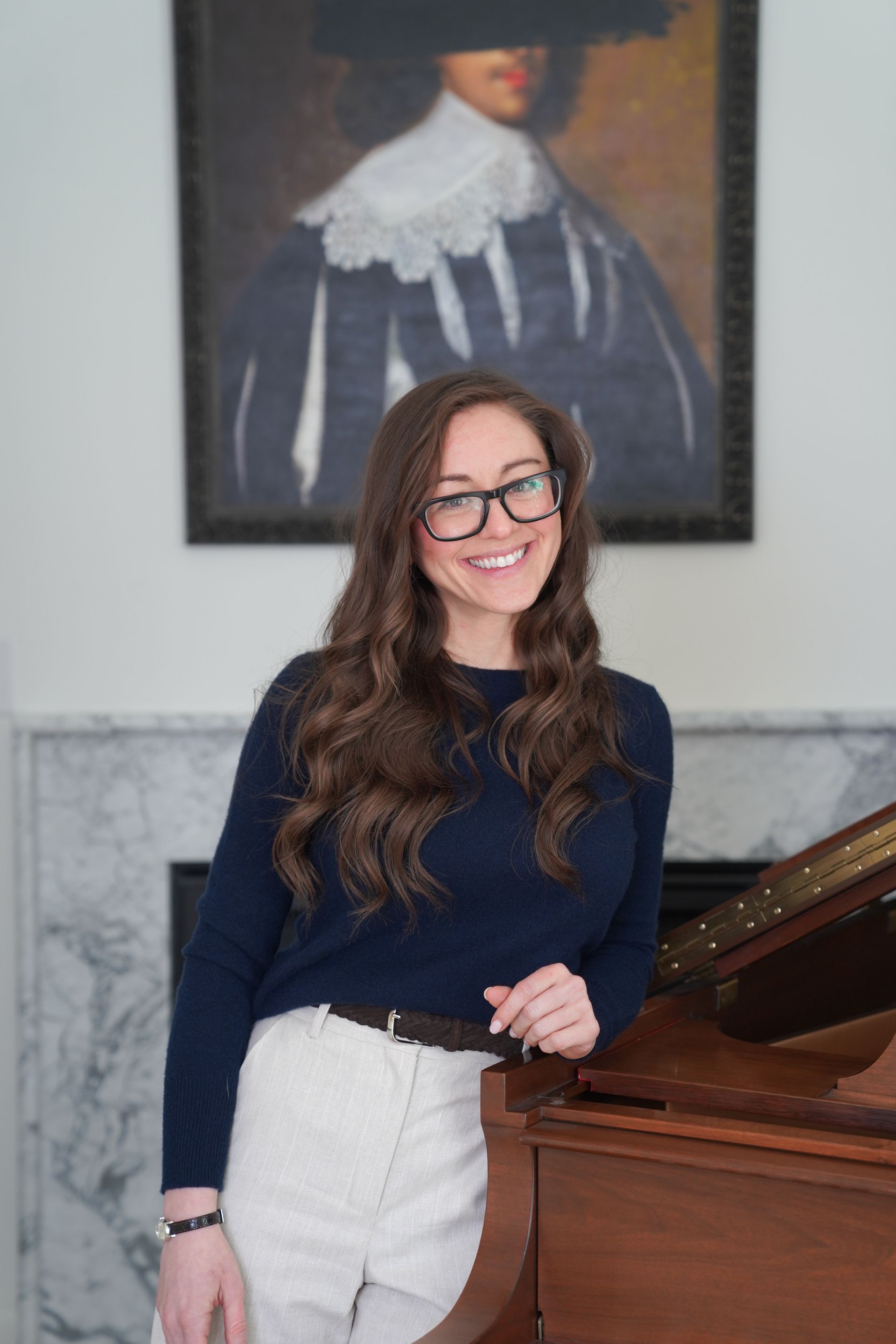Woman in navy sweater and white pants leaning beside a wooden piano under a framed portrait.