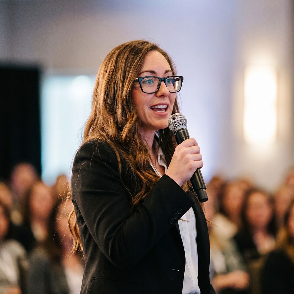 Woman in glasses speaking into a microphone at a conference, with a blurred audience behind her
