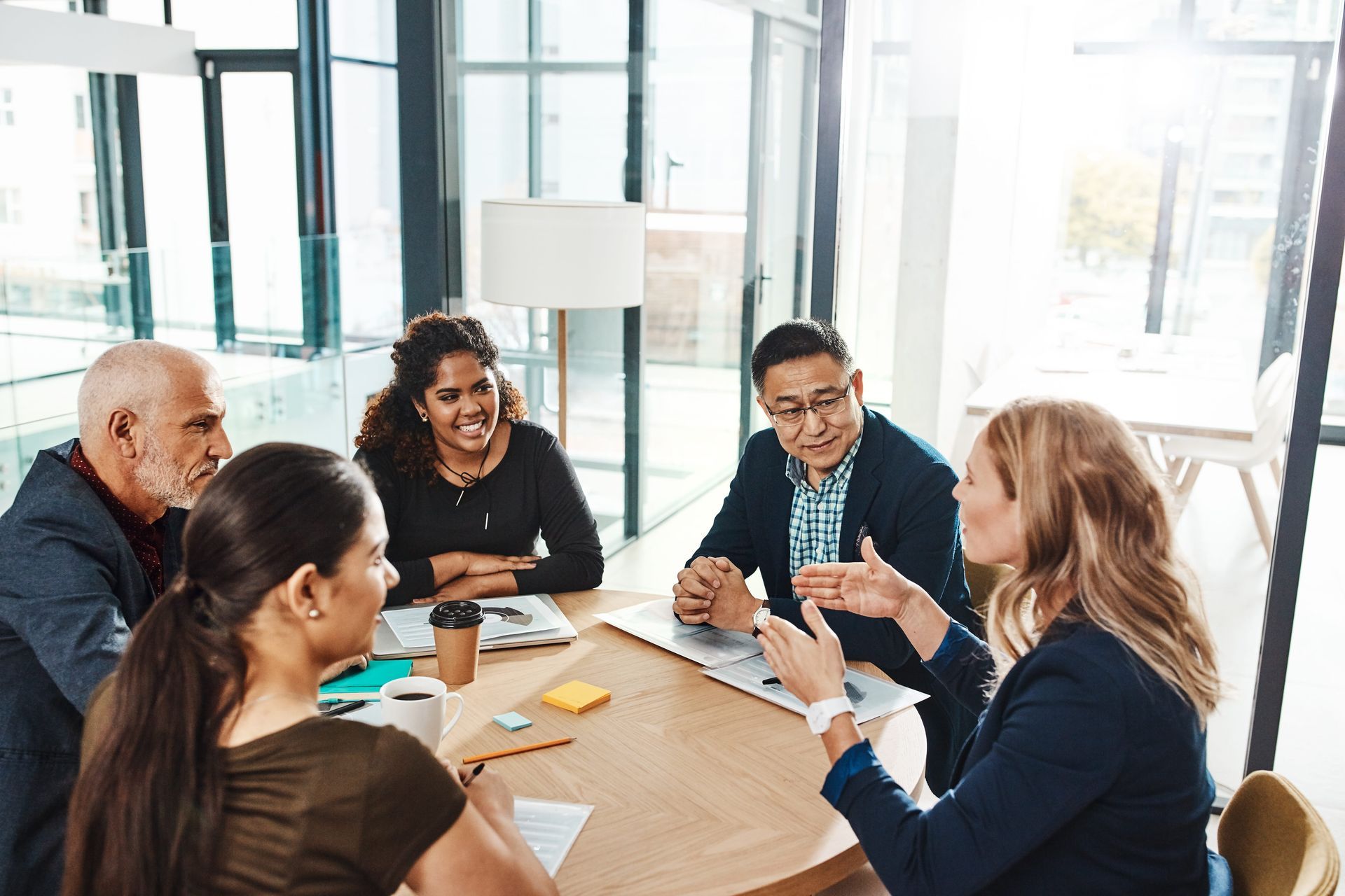Five people in a bright office meeting around a table, talking and taking notes