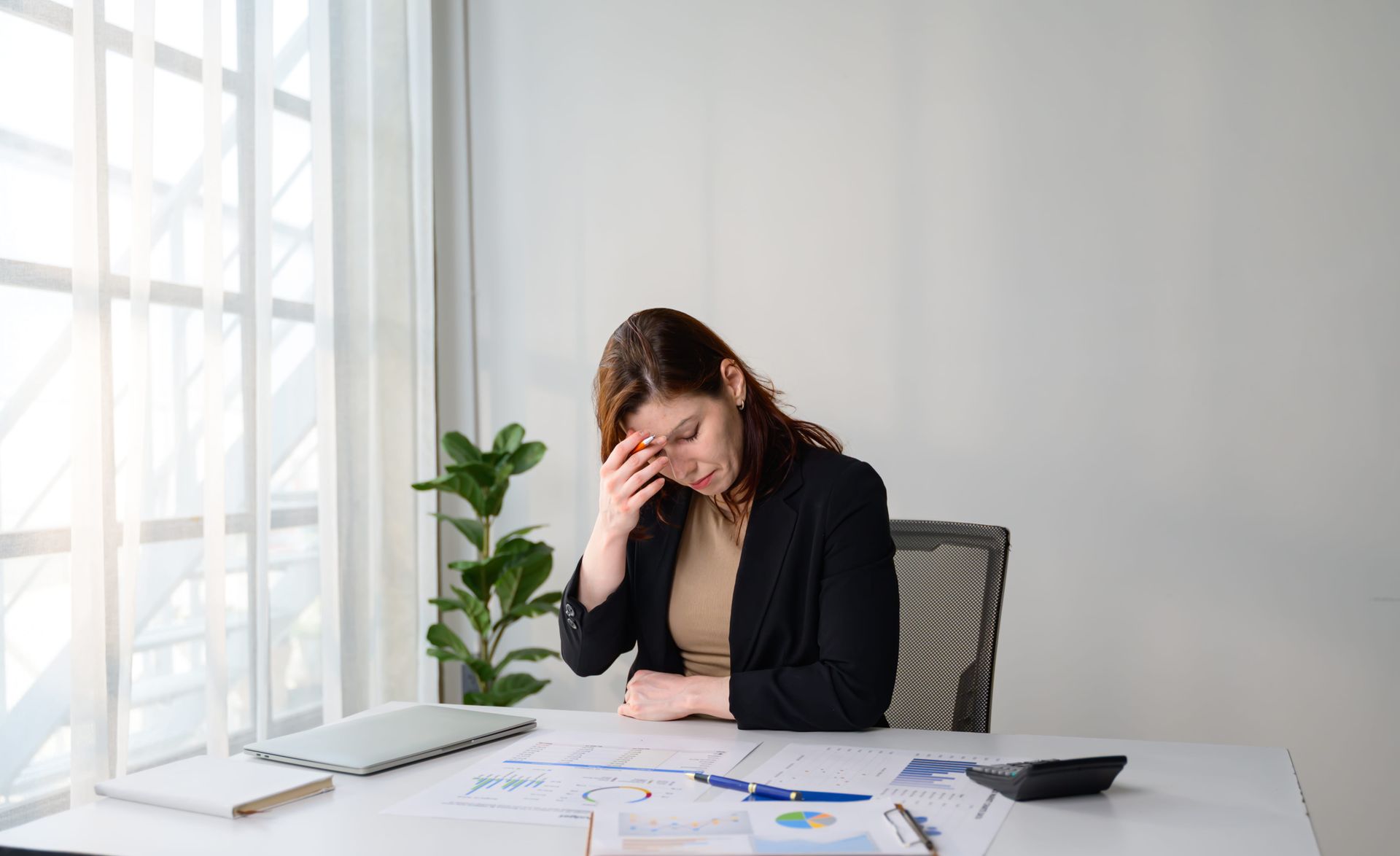 Woman sitting at a desk, holding her forehead, appearing stressed amid scattered papers.
