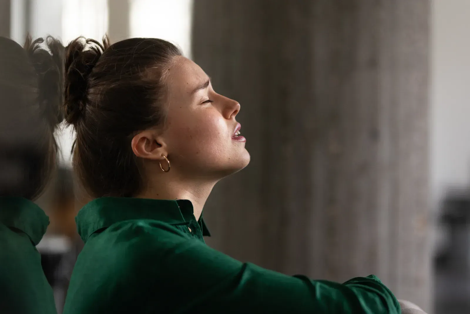 Person in a green top with eyes closed, sitting by a window and facing upward in a calm pose