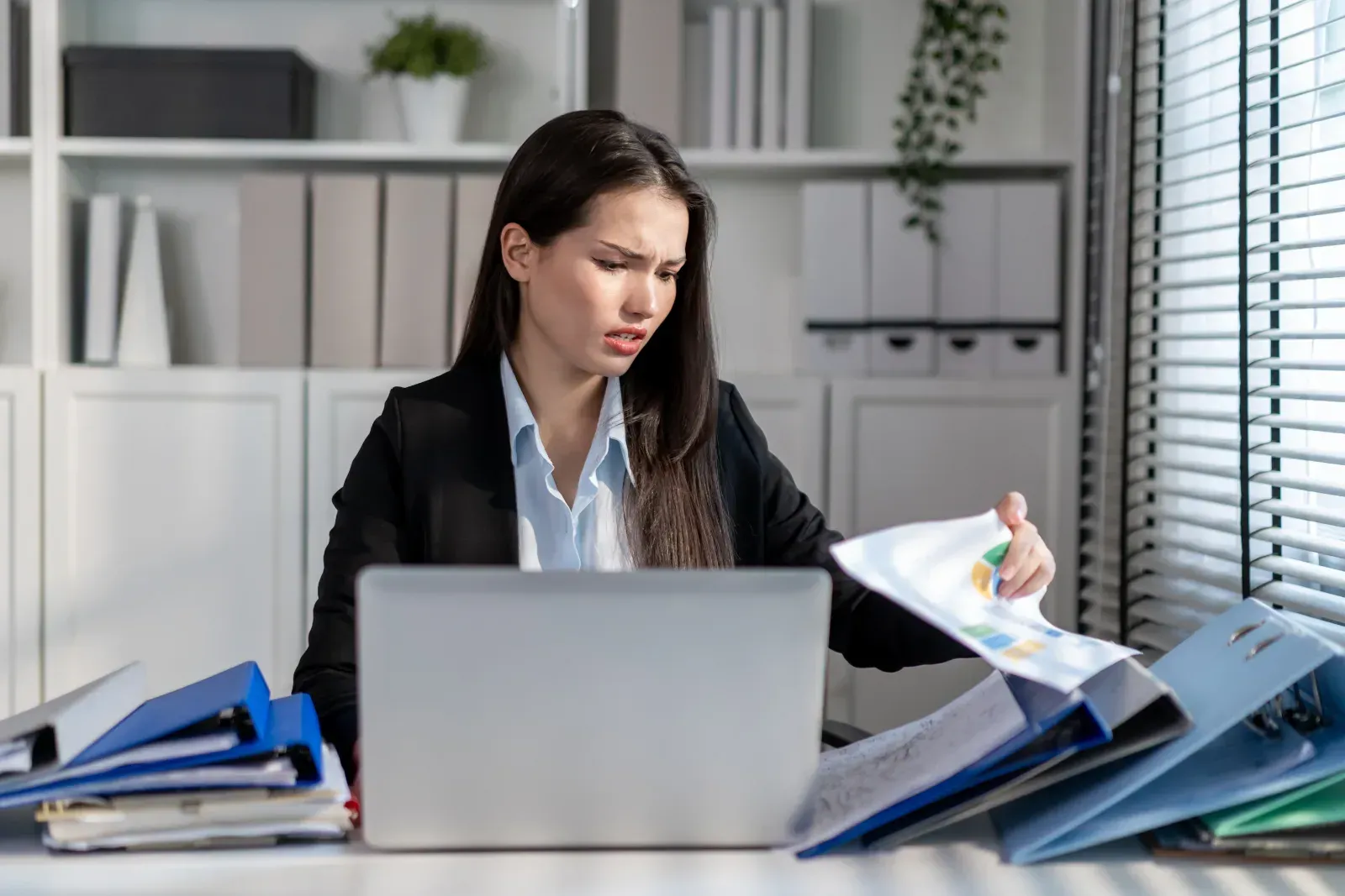 Woman working at desk with laptop and blue folders in a bright office, looking through papers