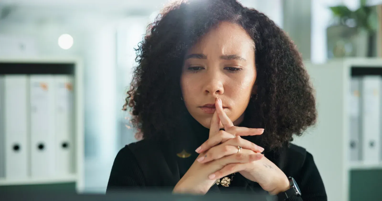 Woman in an office, looking thoughtful with clasped hands at a desk.