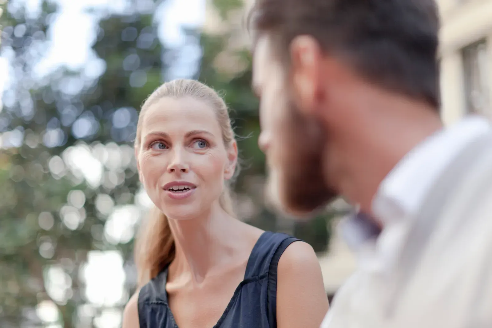 Two people talking outdoors, with a blonde woman in focus and a bearded man blurred in the foreground.
