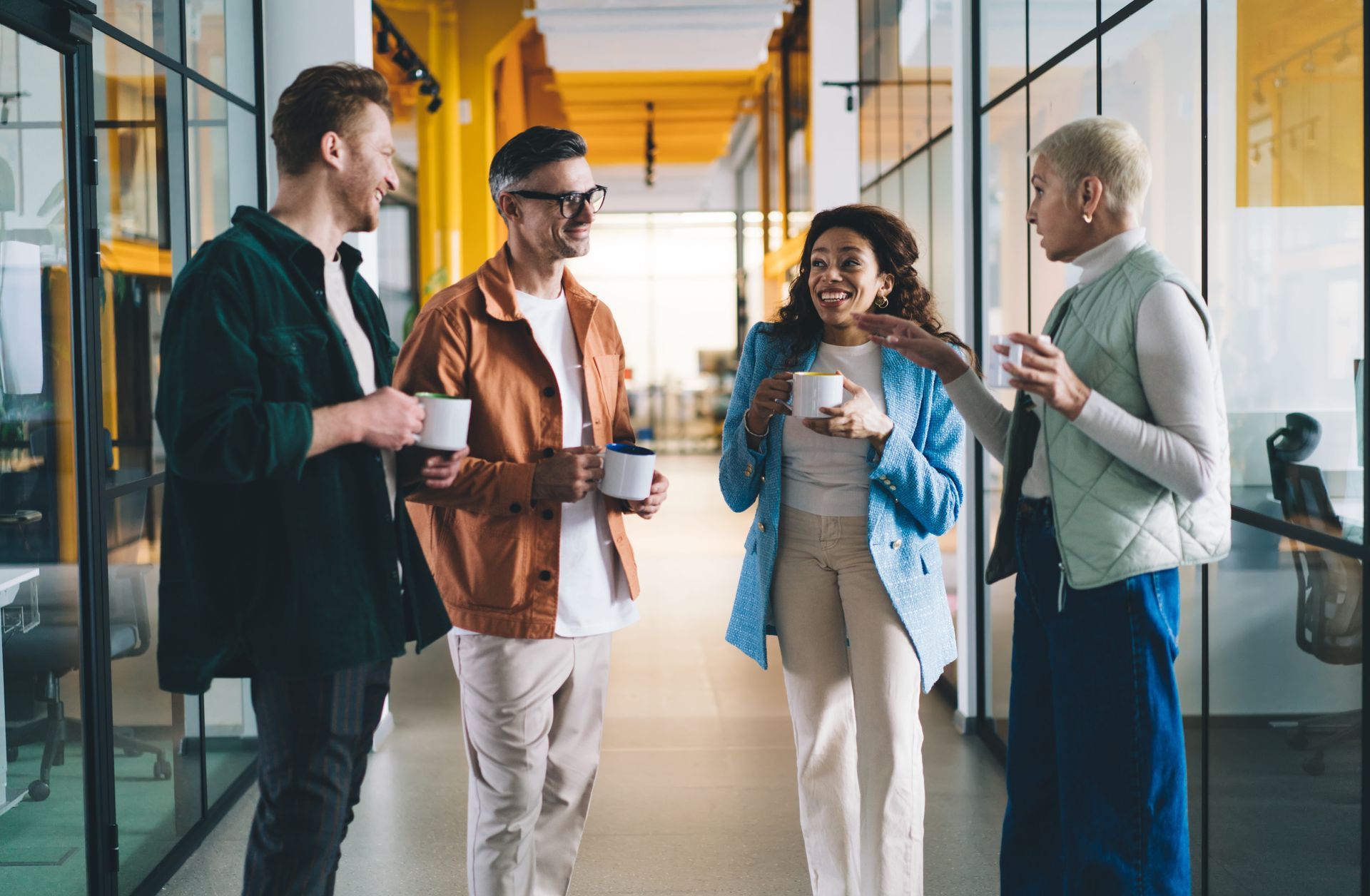 Four colleagues chatting with coffee in a modern office hallway