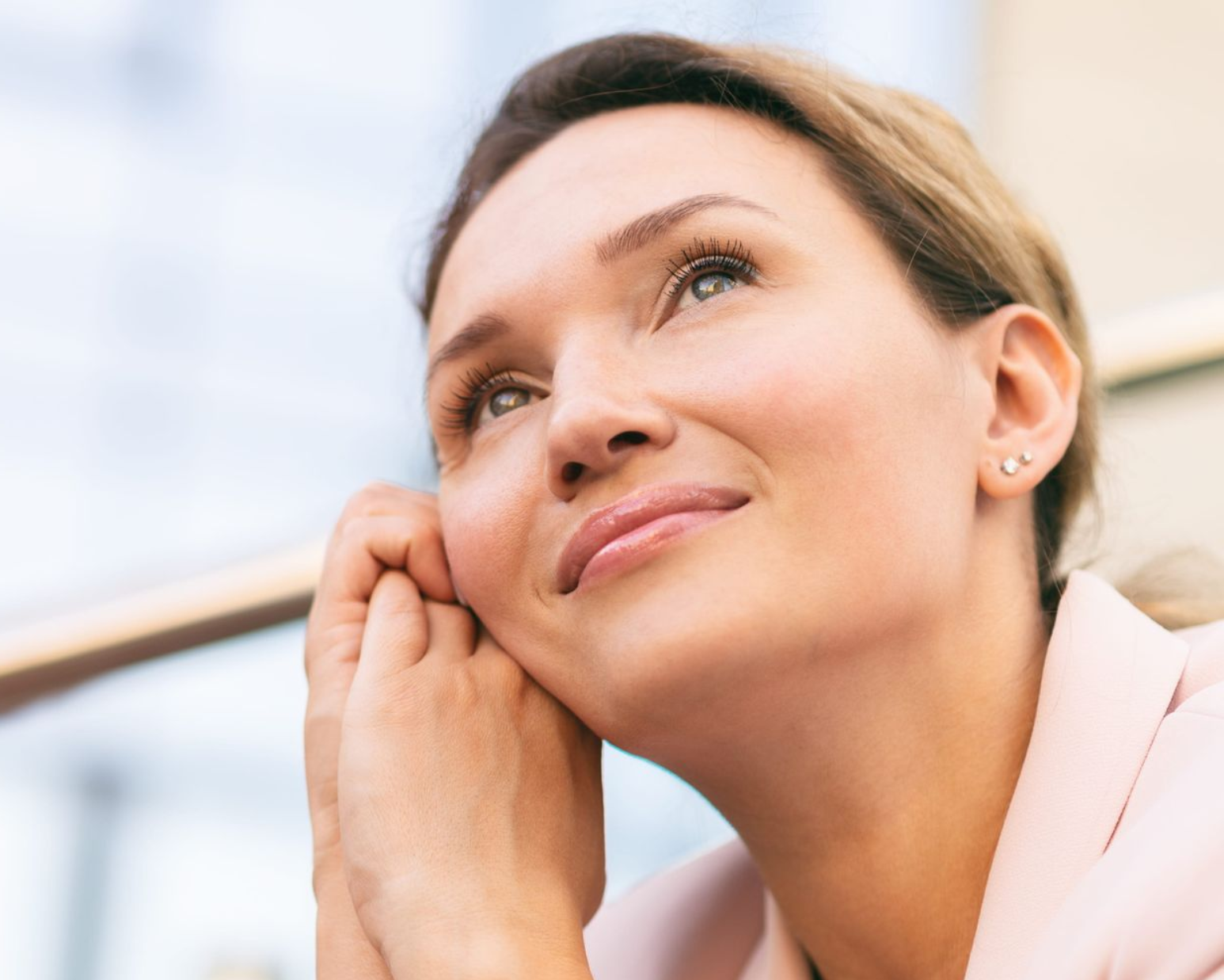 Close-up of a smiling woman gazing upward, resting her hand on her cheek outdoors.