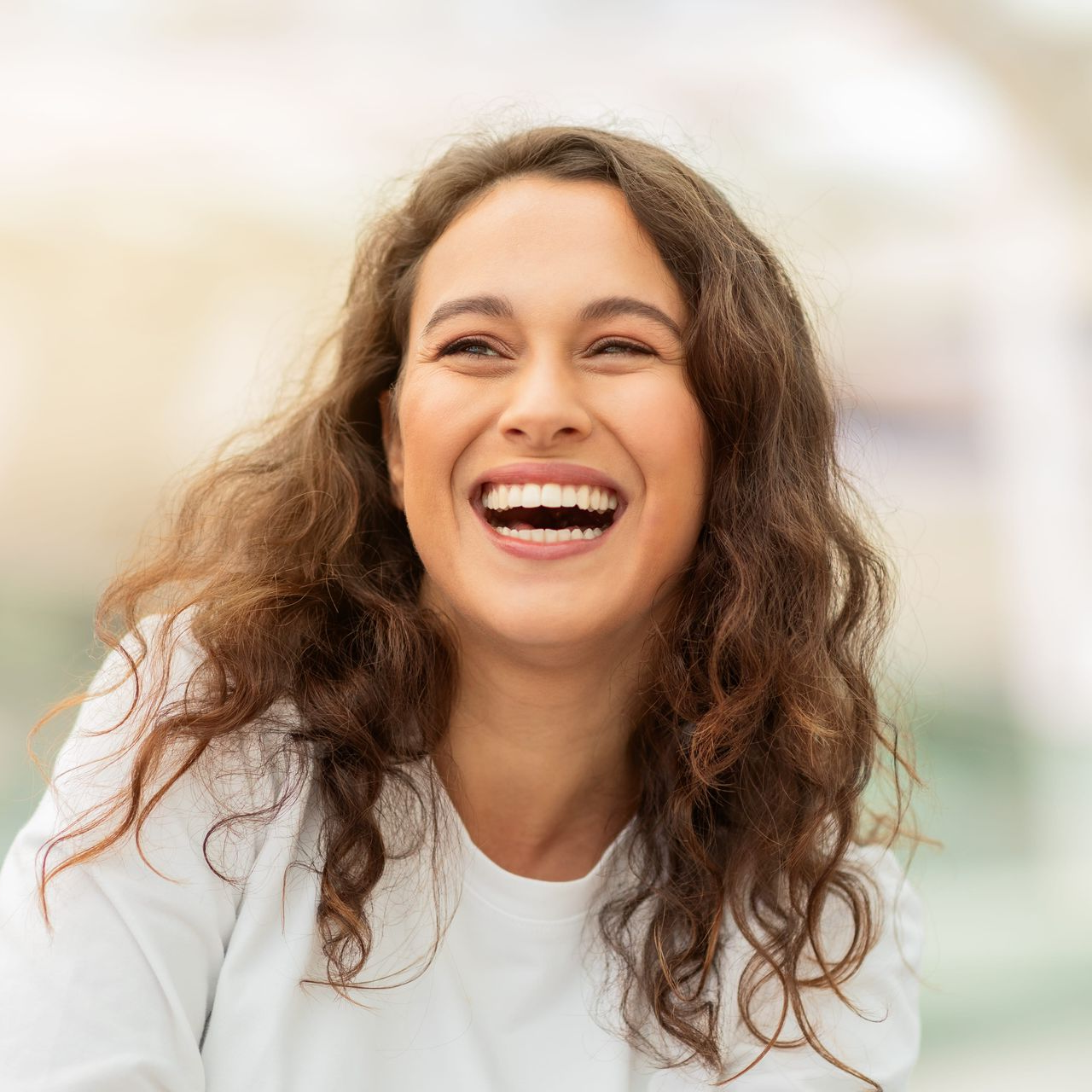 Smiling woman with curly brown hair in a white shirt, laughing against a softly blurred background
