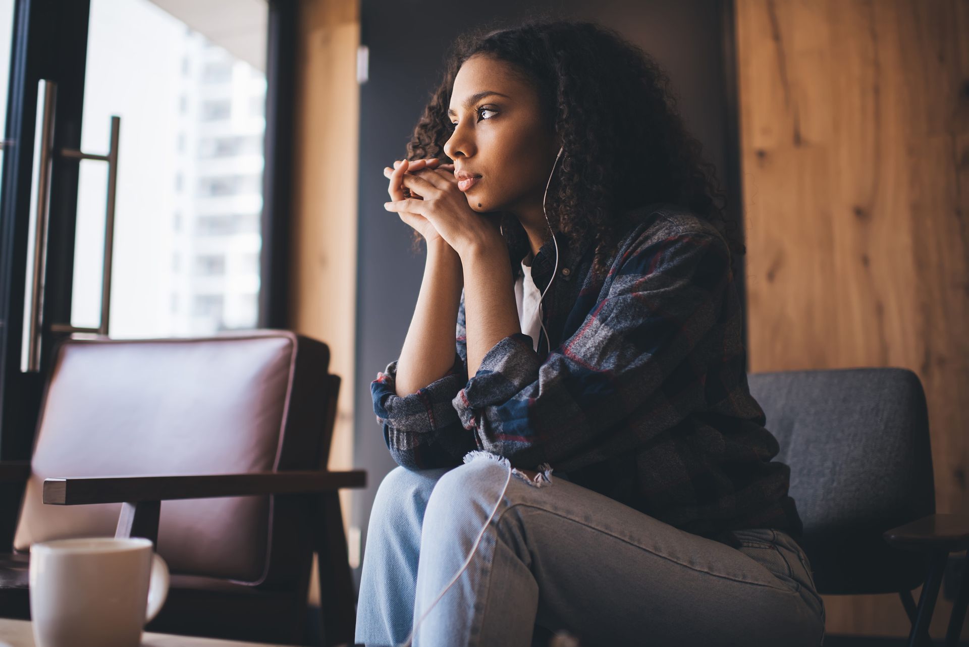 Woman sitting by a window in a cafe, holding a cup and looking outside thoughtfully