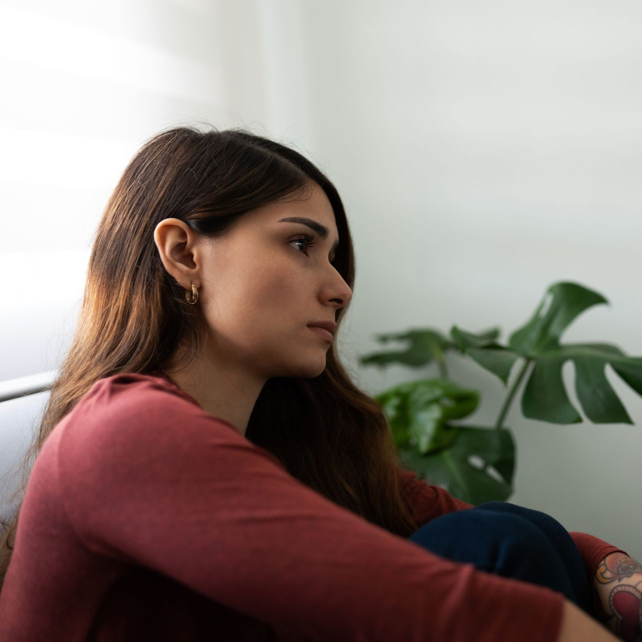 Person sitting indoors by a window, looking away thoughtfully, with a large green plant nearby.
