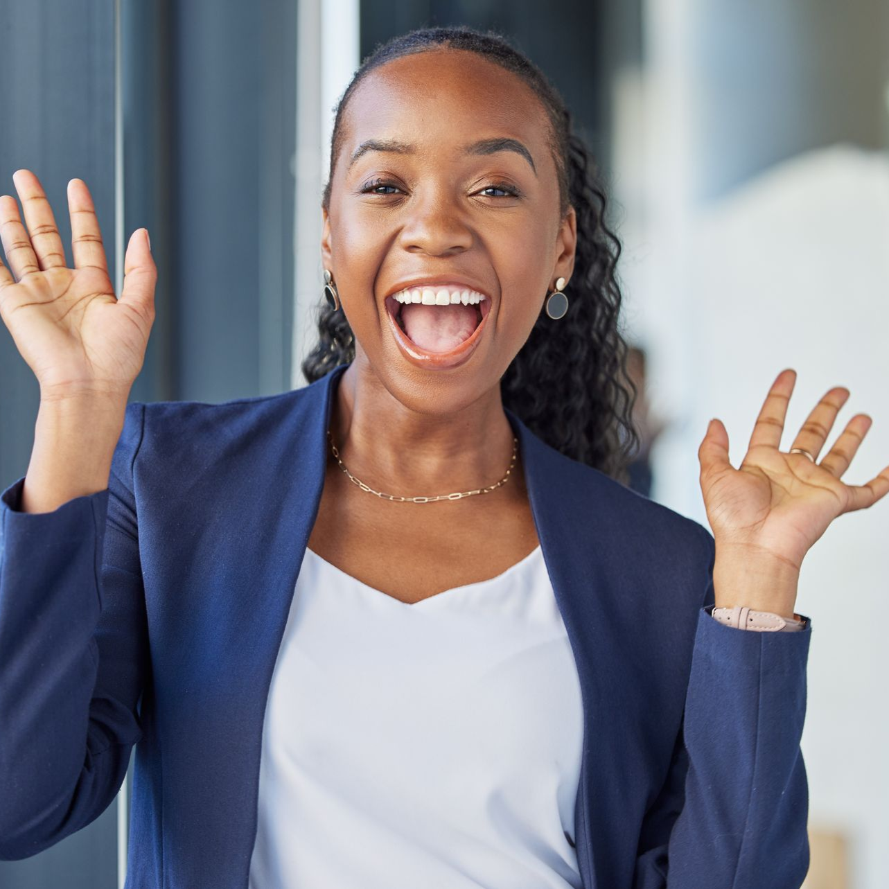 Smiling woman in a blue blazer waving with both hands in a bright indoor setting