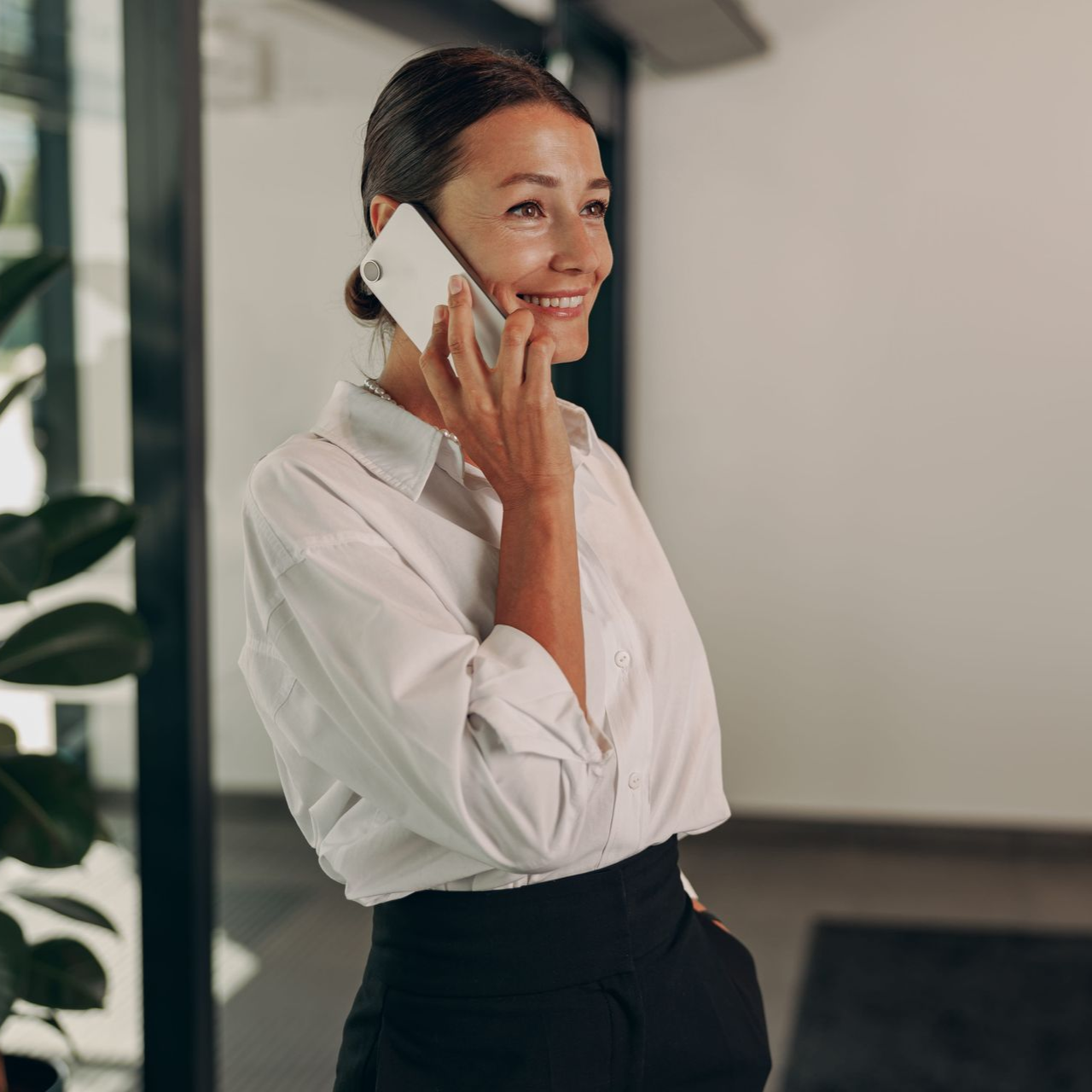 Smiling woman in a white blouse and black skirt talking on a phone in a modern office