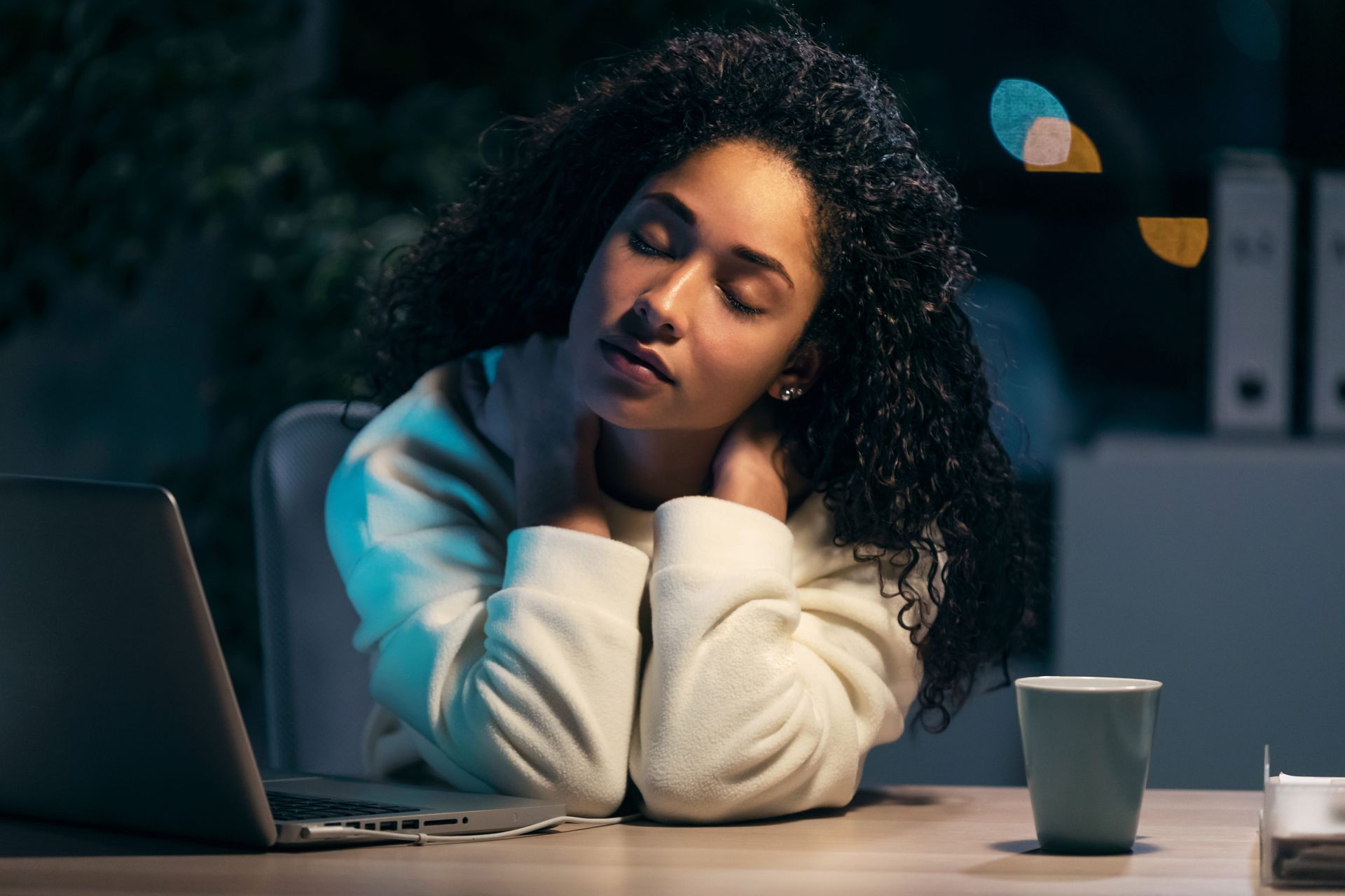 Person resting at a desk beside a laptop and mug, eyes closed in a quiet, dimly lit room