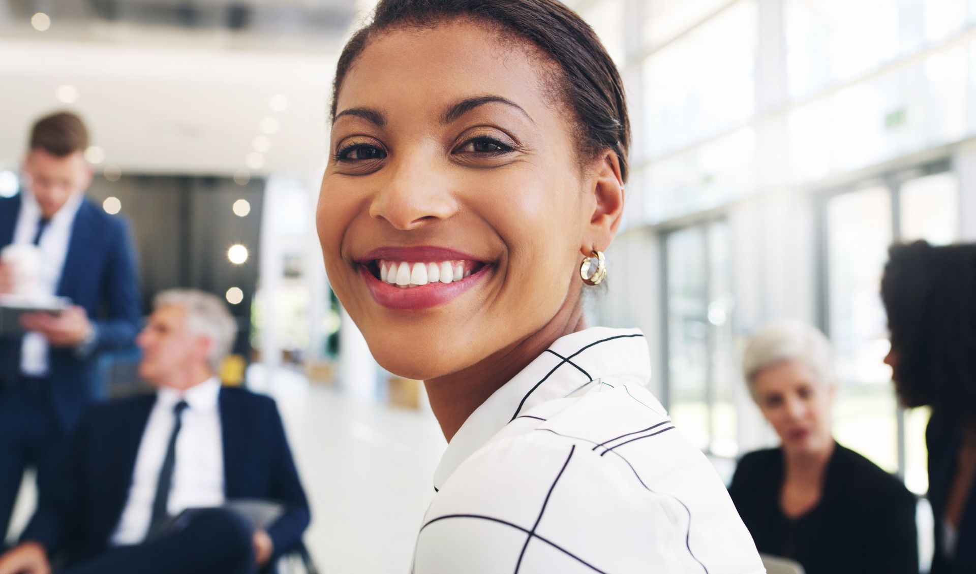 Smiling woman in a white checkered blouse in a bright office with coworkers blurred in the background