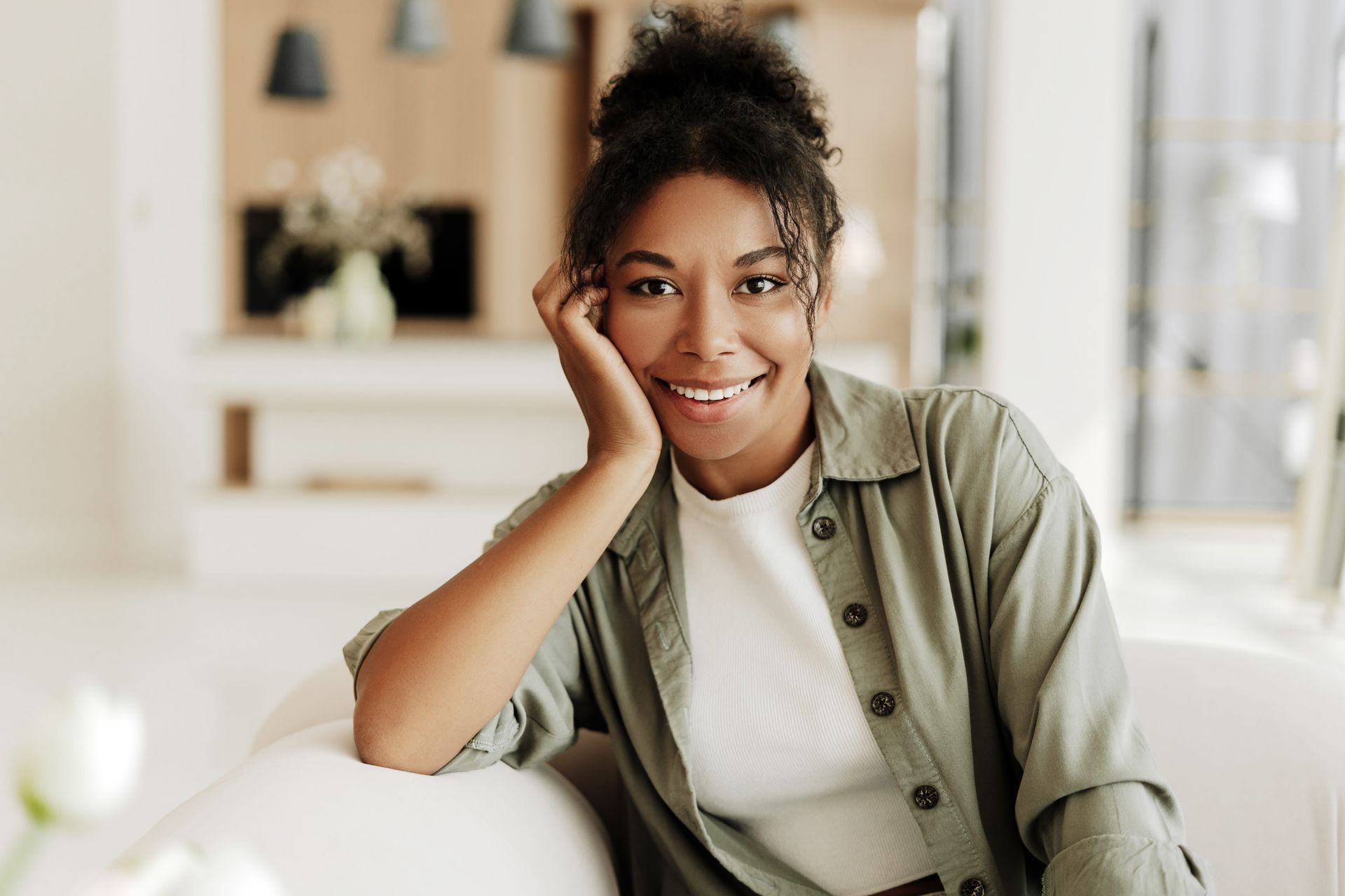 Smiling person sitting on a white couch in a bright living room, resting their head on one hand.