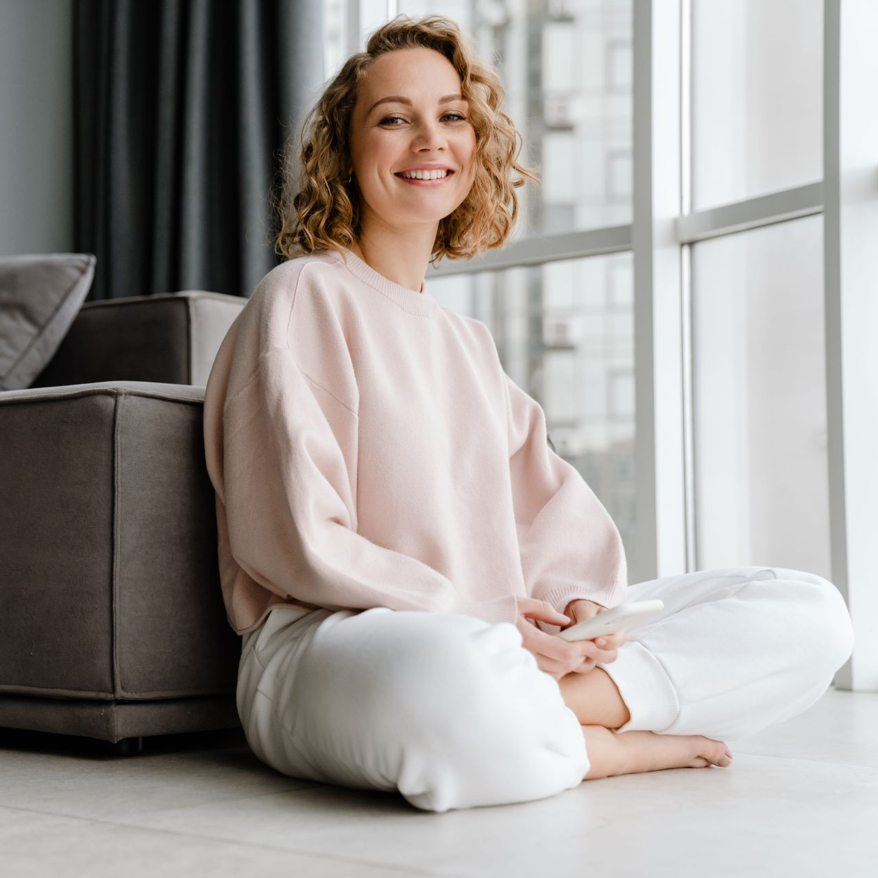 Woman in a light pink sweater sits cross-legged on a floor beside a gray sofa, smiling by a bright window