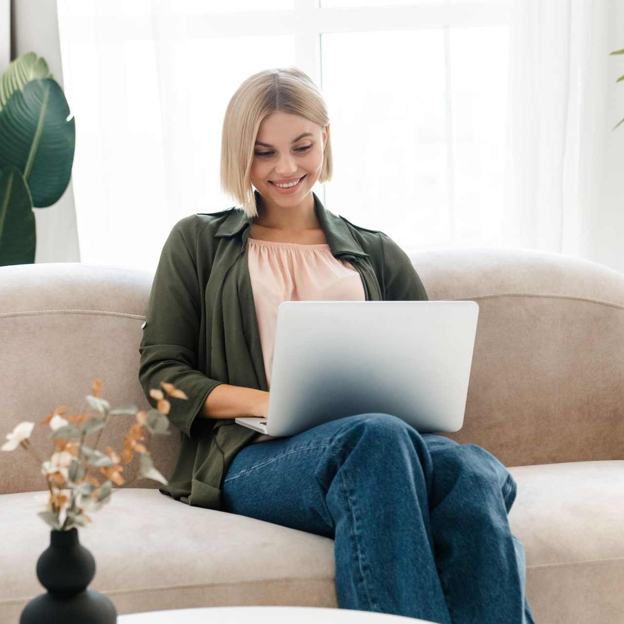 Woman smiling on a beige sofa using a laptop in a bright living room