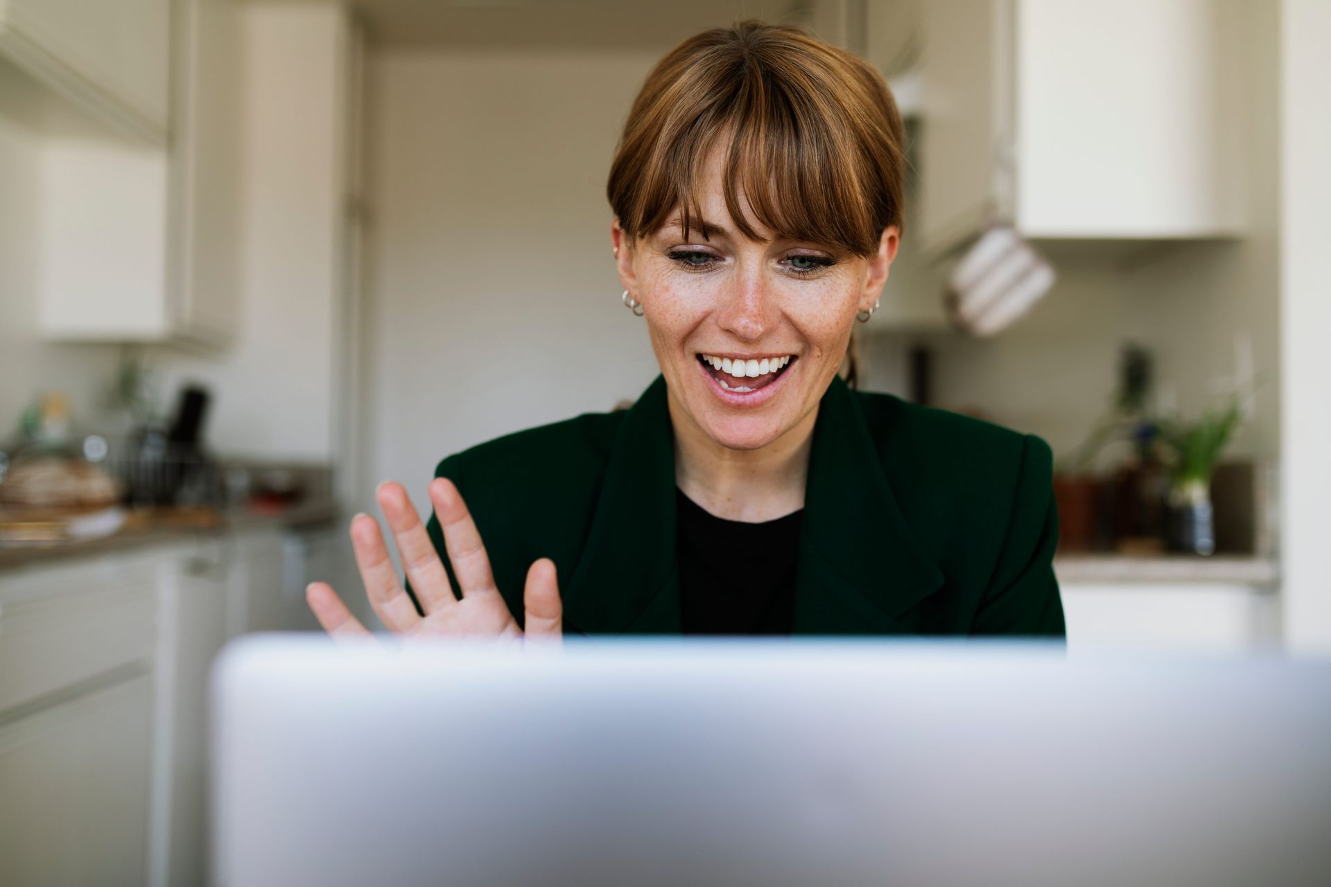 Smiling woman waving on a video call from a kitchen, seen over a laptop screen