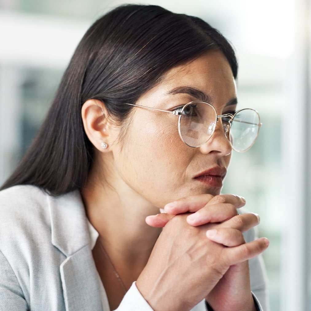 Woman in glasses resting clasped hands under her chin, looking thoughtfully to the side in a bright office