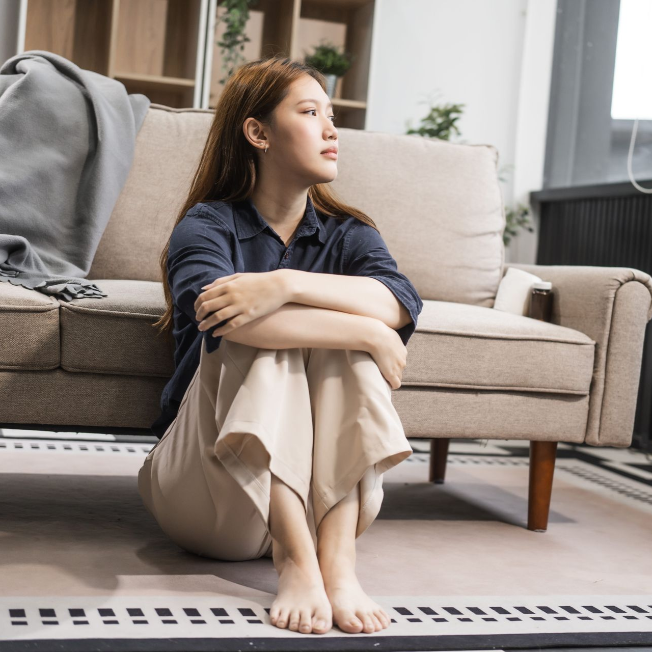 Person sitting on the floor beside a beige sofa, arms wrapped around knees, looking out a window.