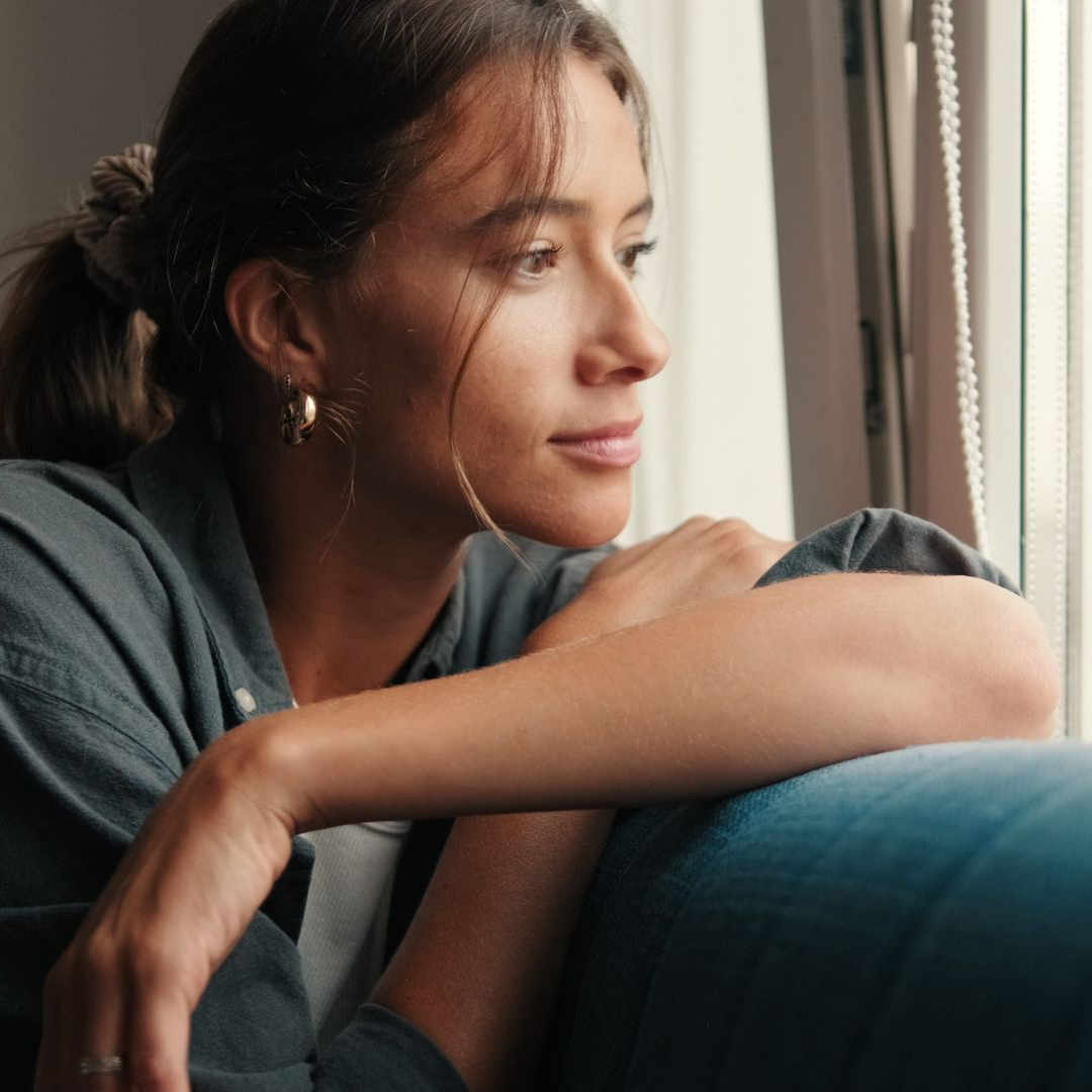 Young woman gazing out a window, resting her arms on a blue couch