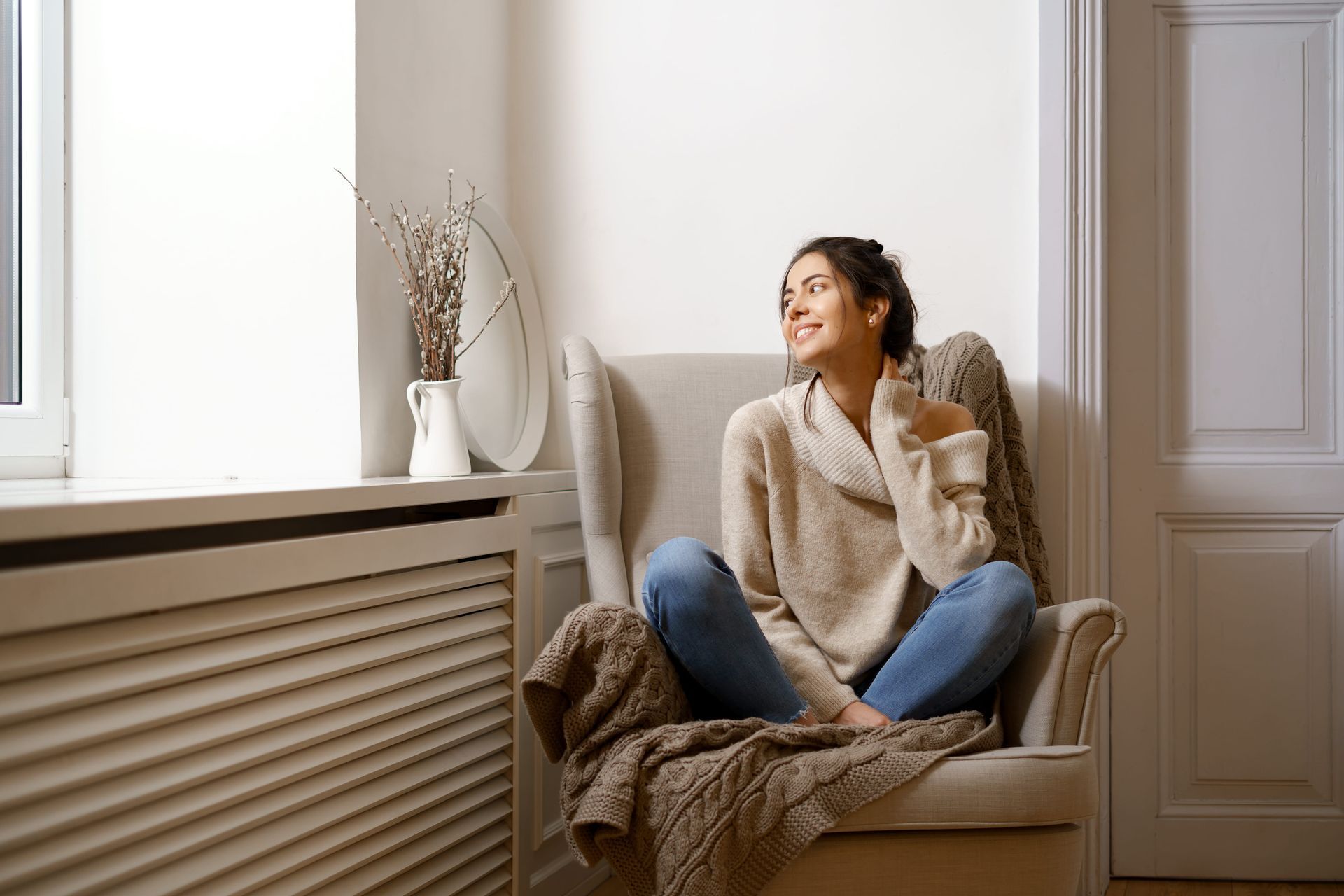 Woman sitting in a cozy armchair by a bright window, smiling and relaxing with a cup in hand