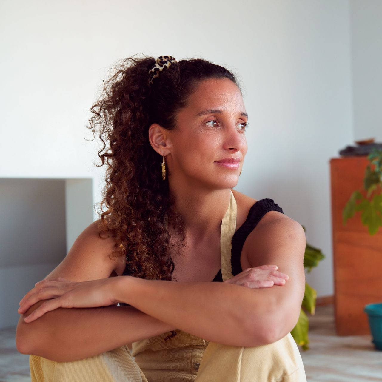 Woman sitting with arms crossed, looking to the side in a bright room with a potted plant.