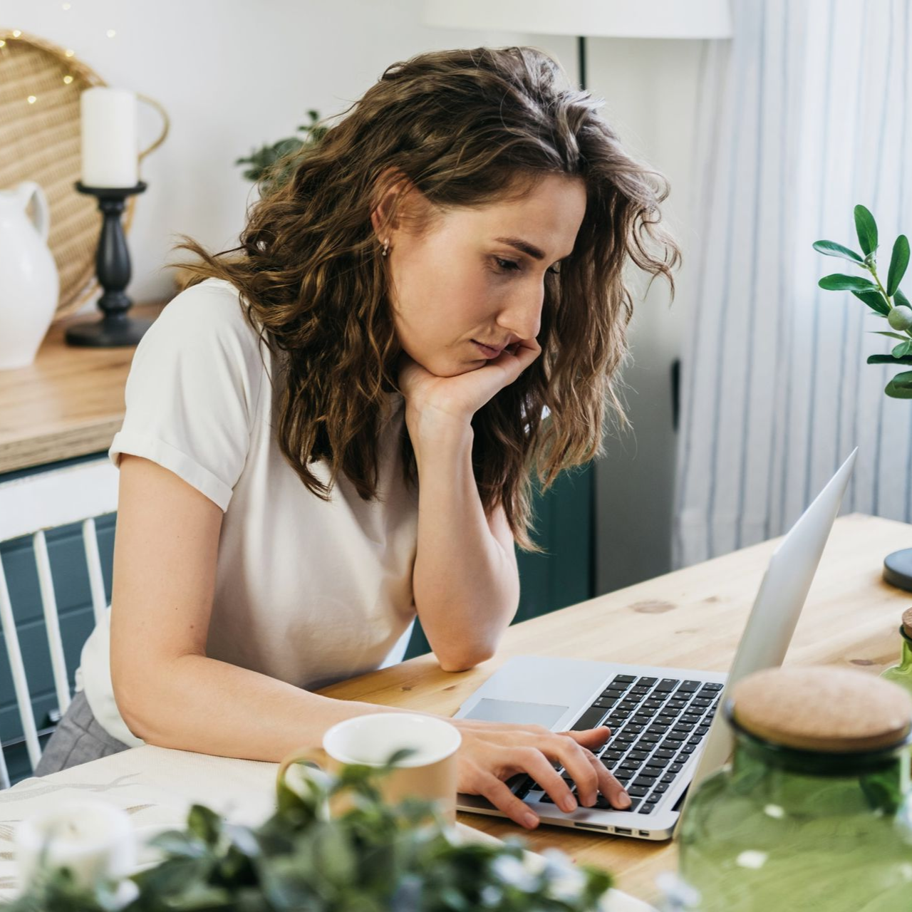 Woman working on a laptop at a kitchen table, leaning on her hand, with a coffee cup and plants nearby