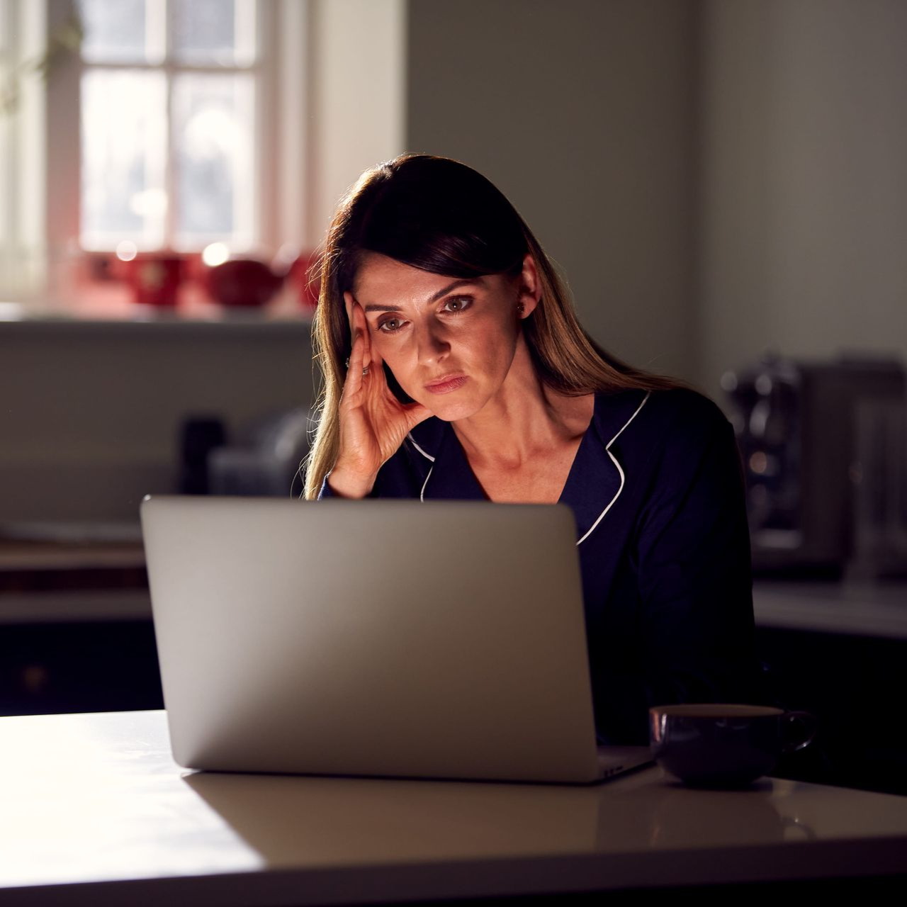 Woman studying at a laptop in a dim kitchen, resting her head on her hand.
