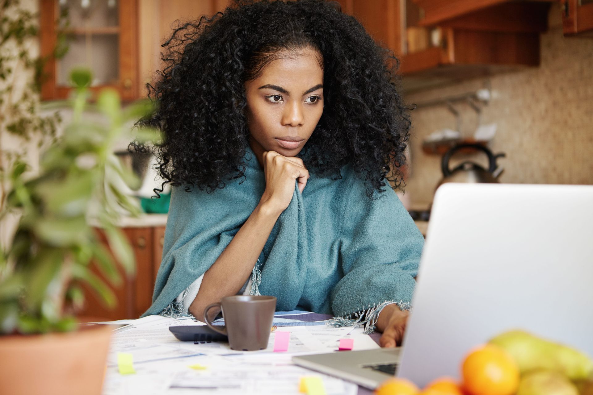 Woman studying at a table with papers and a laptop, resting her chin on her hand in a home kitchen.