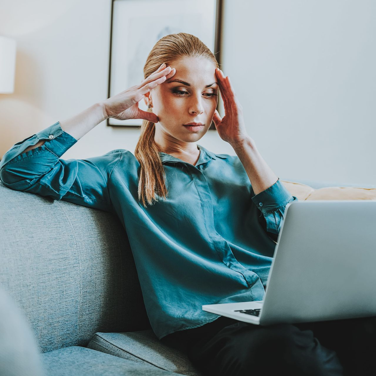 Woman in teal sweater holding her temples while using a laptop on a couch