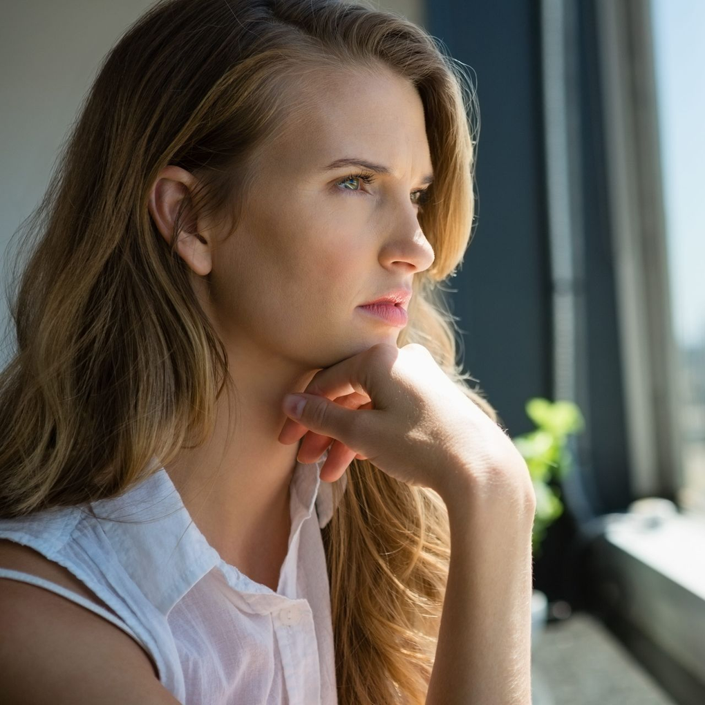 Woman in a white shirt gazing out a window, resting her chin on her hand, in soft sunlight