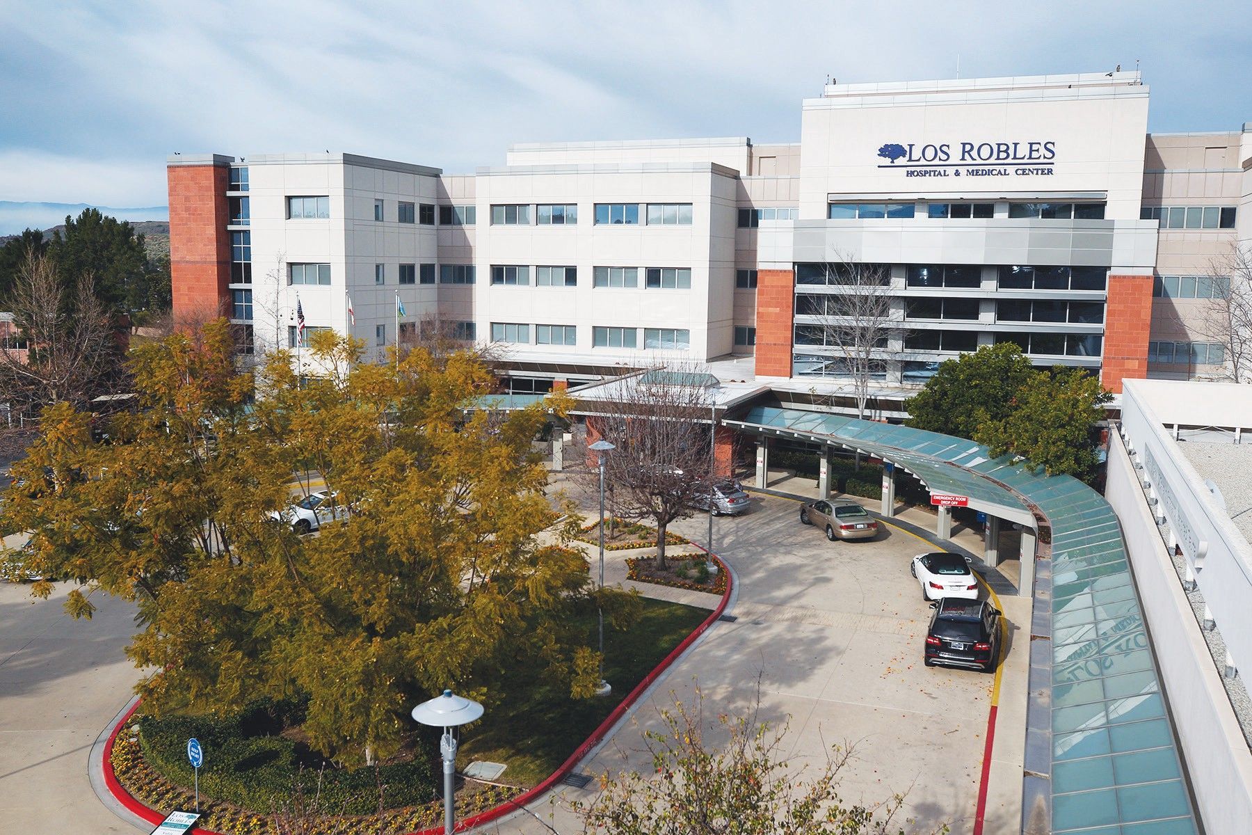 Hospital building with curved glass entrance, parked cars, and autumn trees in front