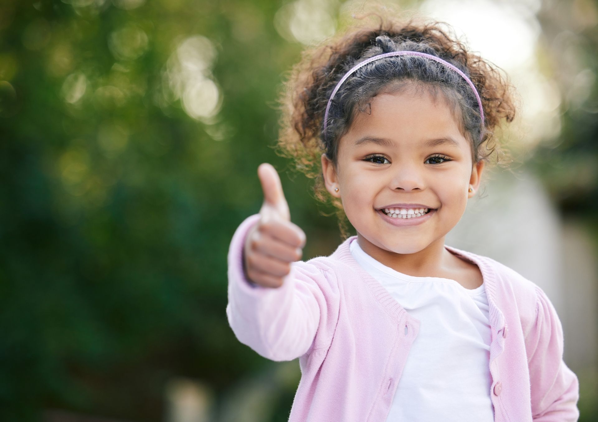 Young girl with curly hair smiles, giving a thumbs-up in an outdoor setting.