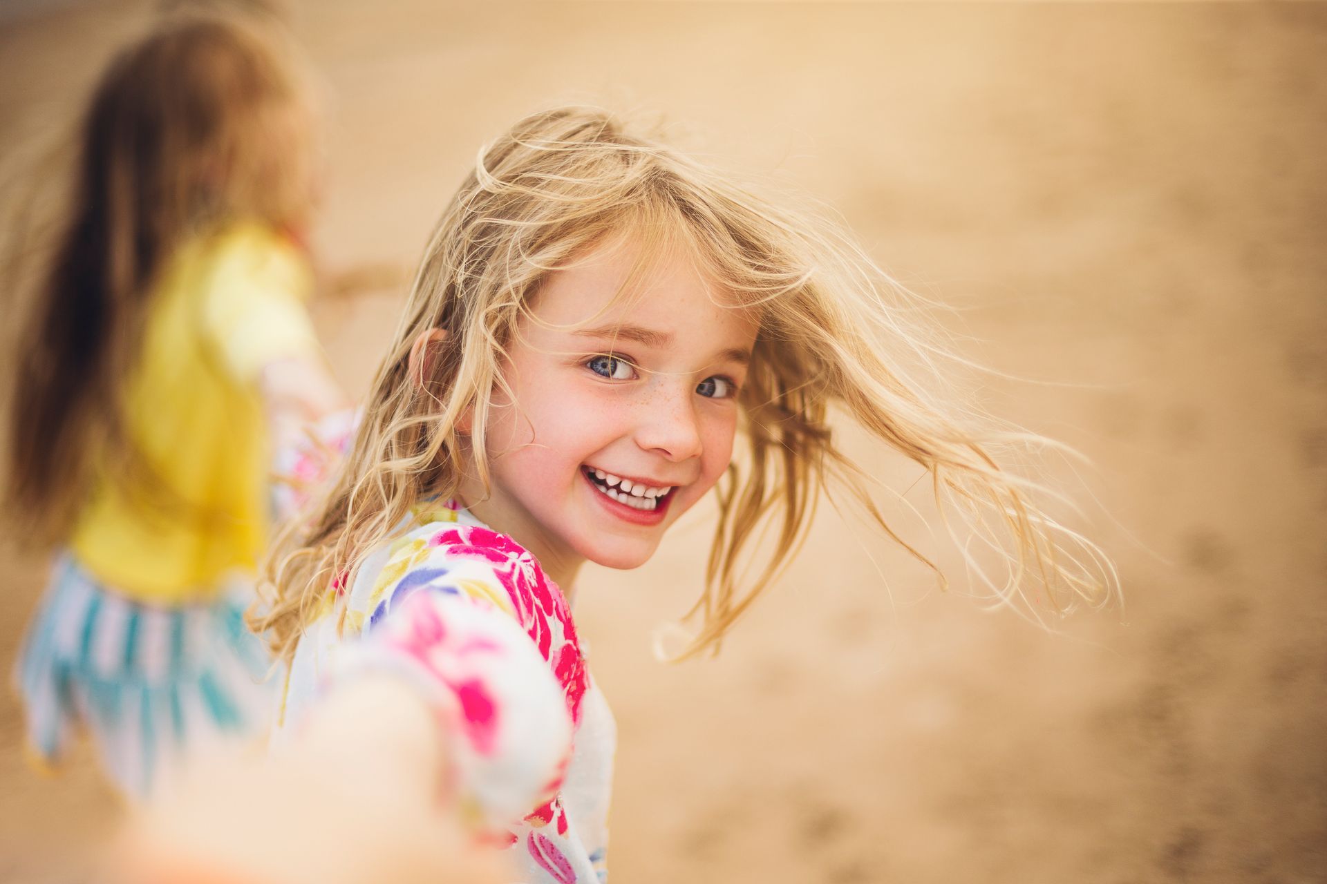 Blonde girl smiles, looking back at the camera. She's holding someone's hand on a sandy beach.