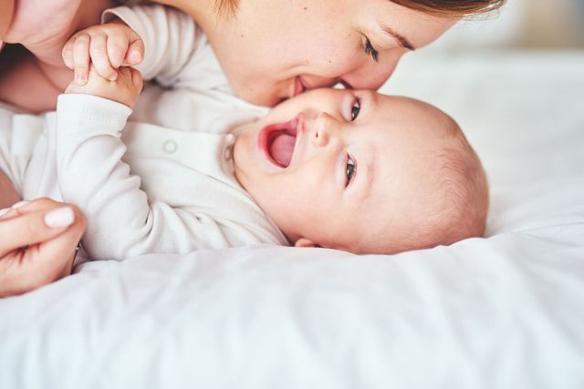 A mother tenderly kisses a smiling baby lying on a white bed.