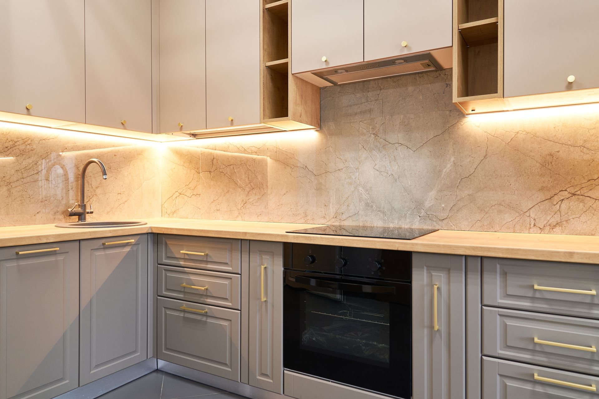 Modern beige kitchen with gray cabinets, marble backsplash, under-cabinet lighting, sink, and built-in oven