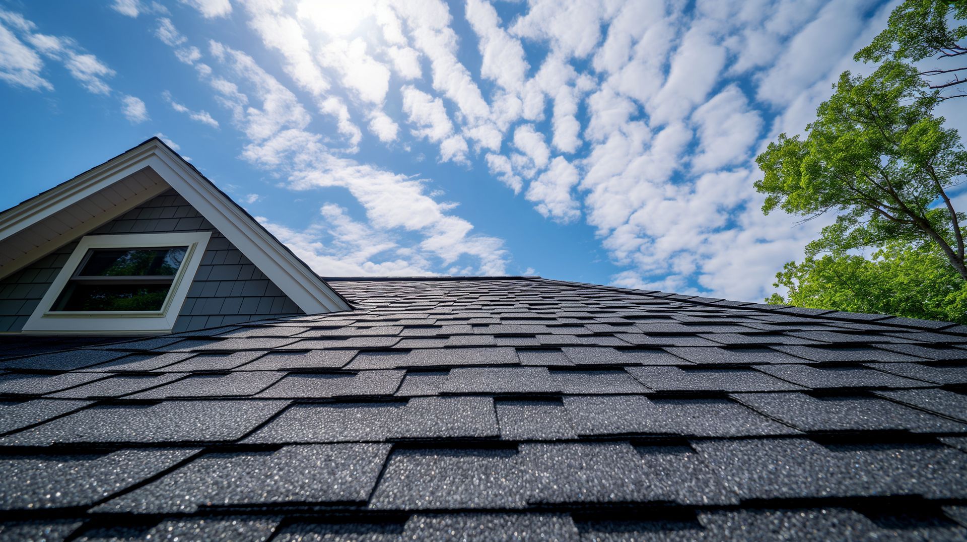 Close-up of a gray shingled roof with a dormer window under a blue sky with scattered clouds