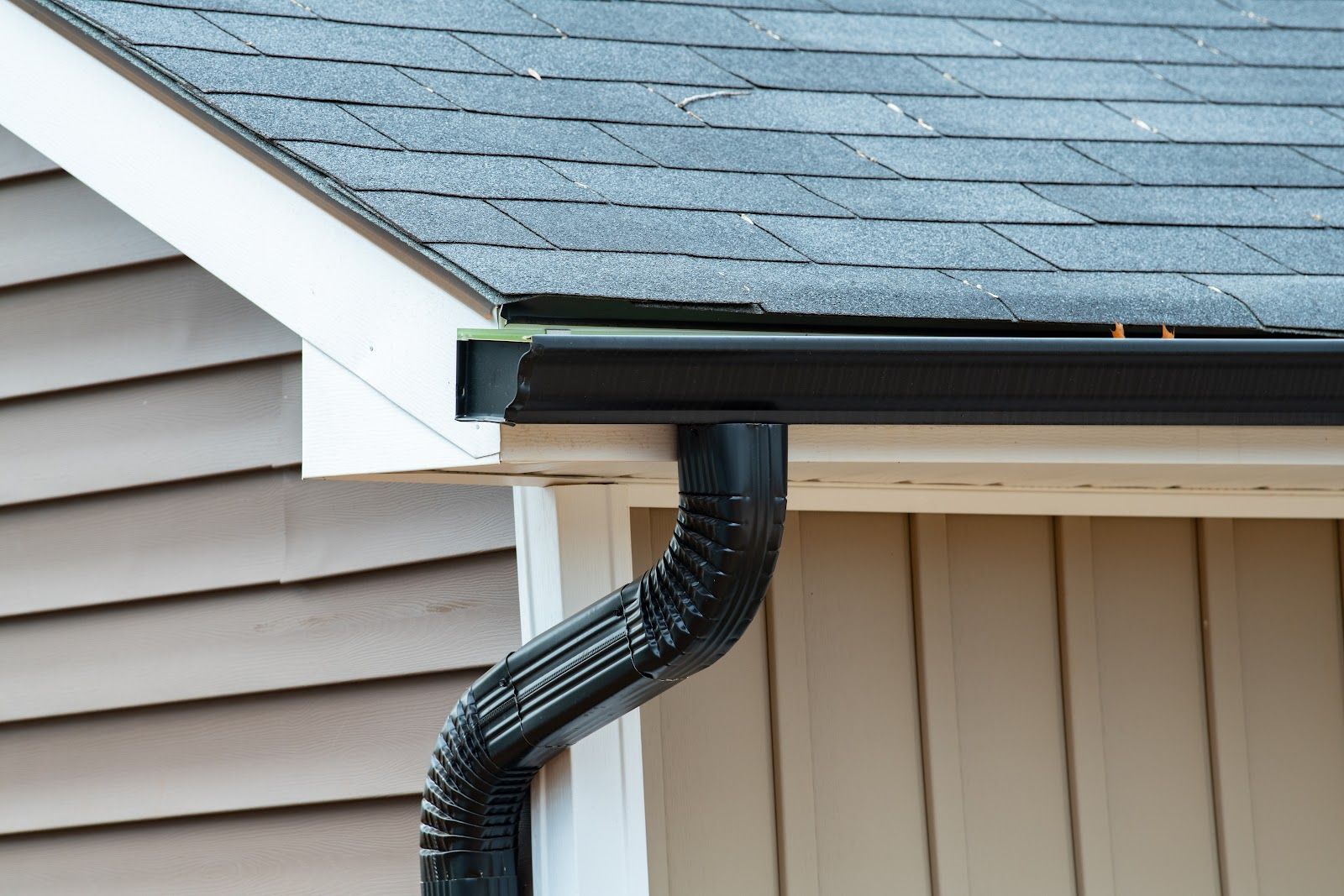 Black rain gutter attached to a tan building with gray roof shingles.