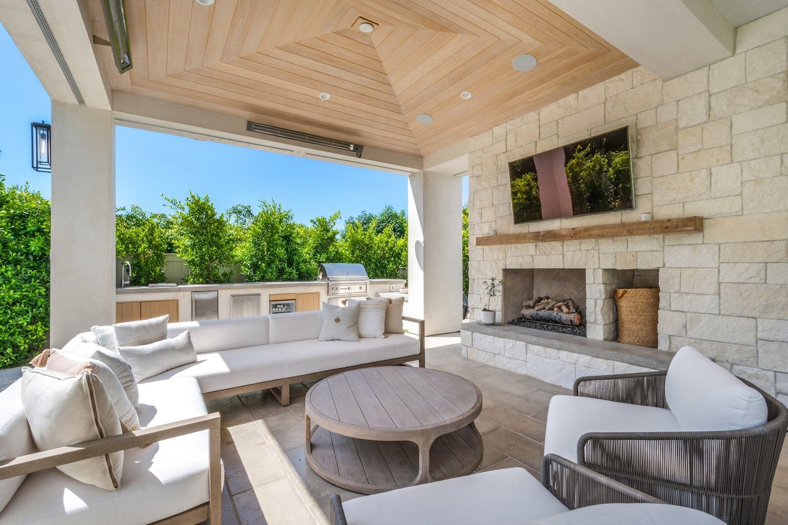 Outdoor living space with white couches, fireplace, TV, and grill.