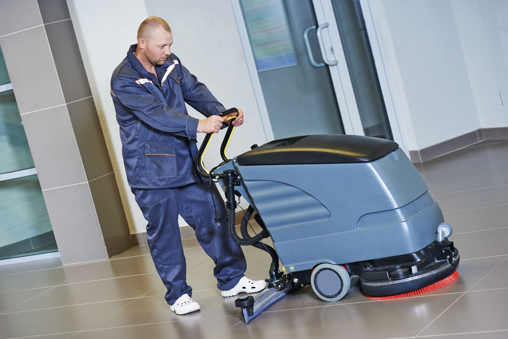 Male worker, wearing a navy blue uniform, cleans the floor of a hall with a washing machine.