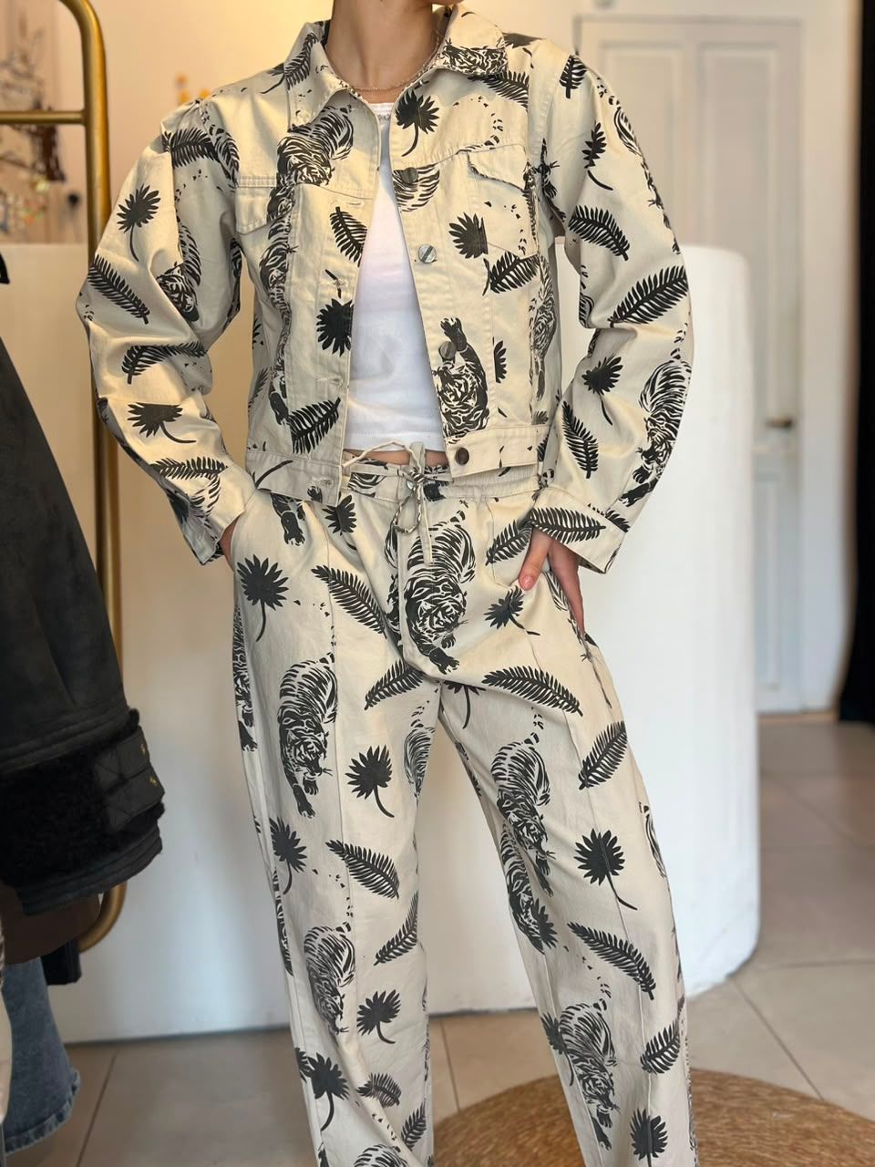 Persona con chaqueta estampada de flores color crema y pantalones a juego, de pie con las manos en los bolsillos en el interior de un edificio.