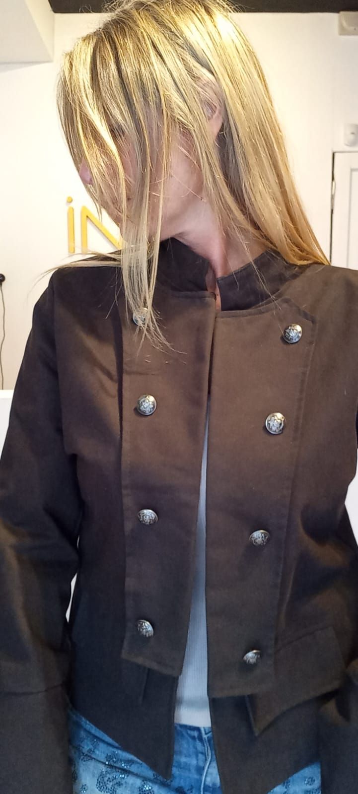 Persona con chaqueta beige y vaqueros azules, de pie en el interior de un edificio, de perfil, mirando hacia un lado.