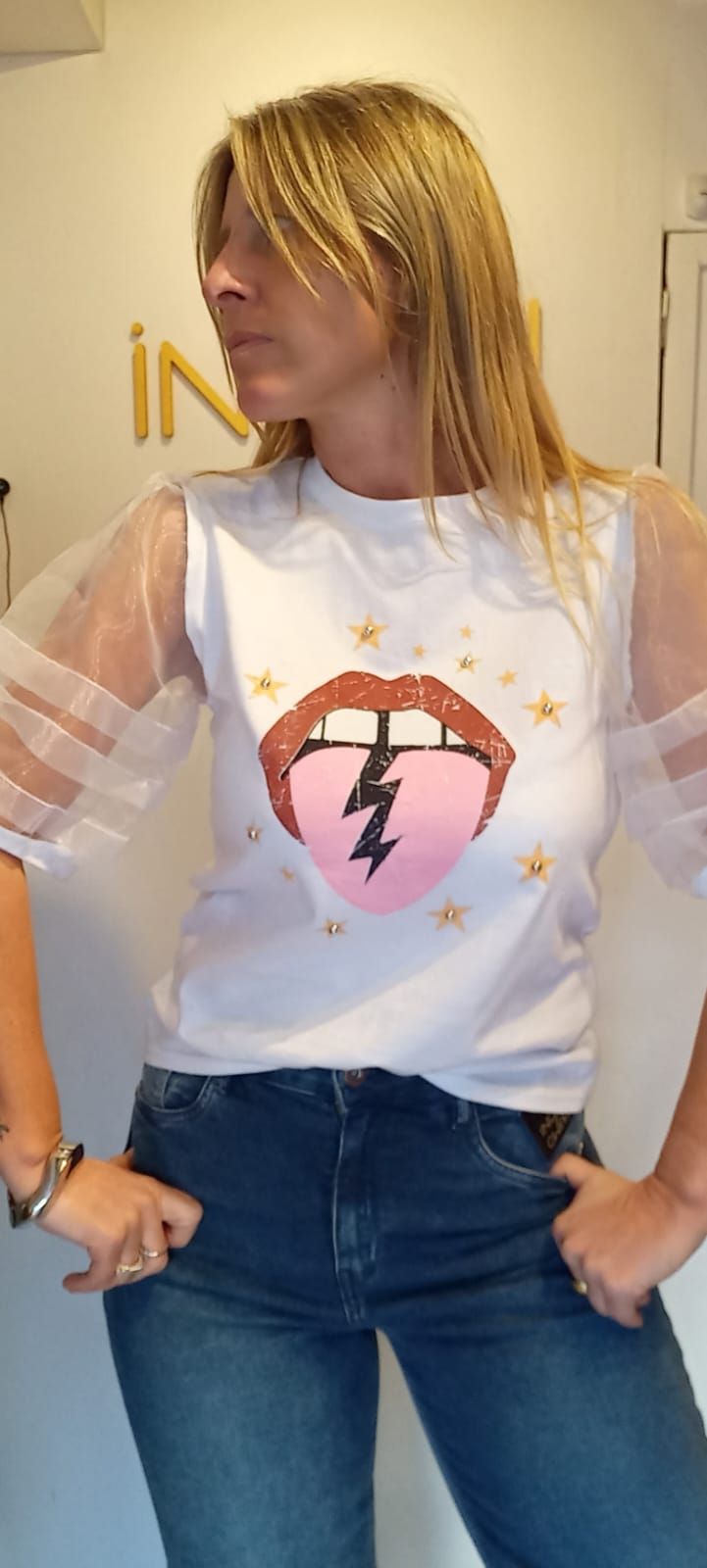 Persona vestida con vaqueros y una camiseta blanca estampada, de pie en el interior de un edificio, con las manos en las caderas y mirando hacia un lado.