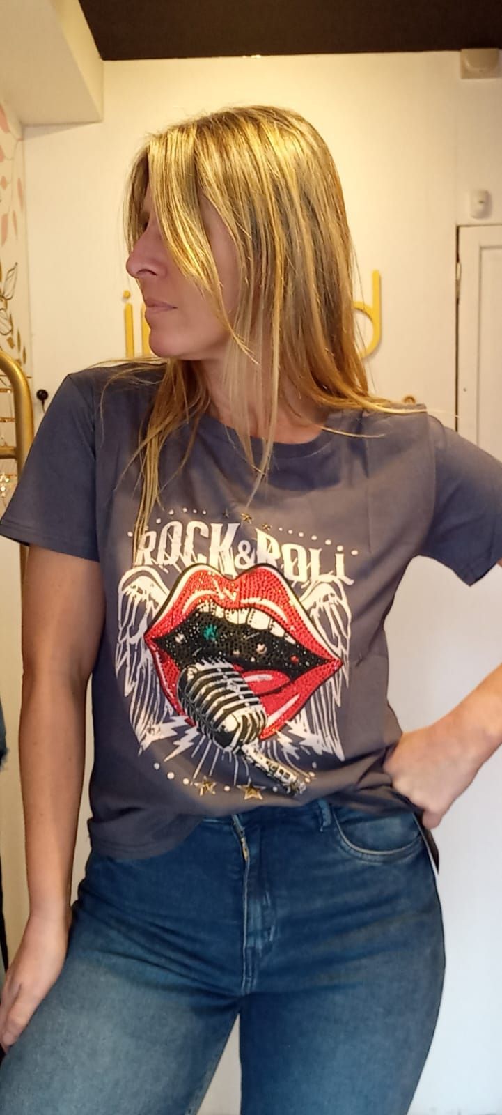 Persona con camiseta azul estampada y vaqueros posando en interiores frente a una pared clara.